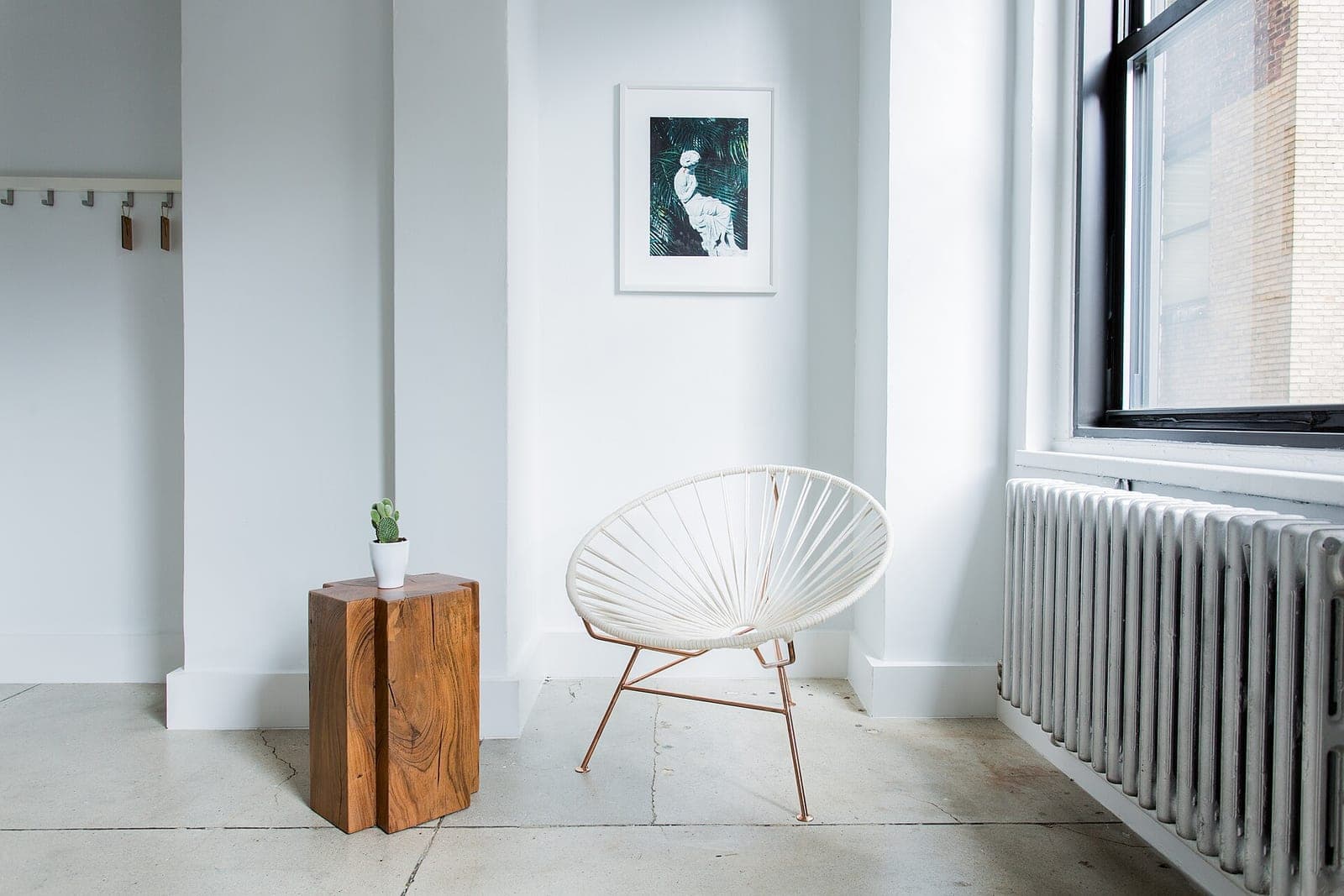 A white woven chair, a wooden side table with a small cactus and a framed print in a bright corner beside a window