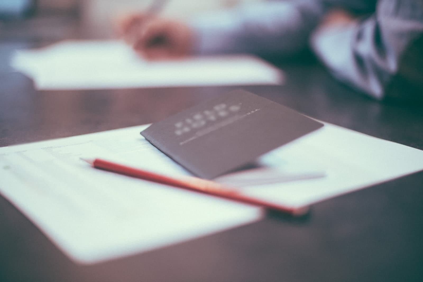 A pencil and a small notebook rest on printed pages on a meeting table while someone across the table takes notes
