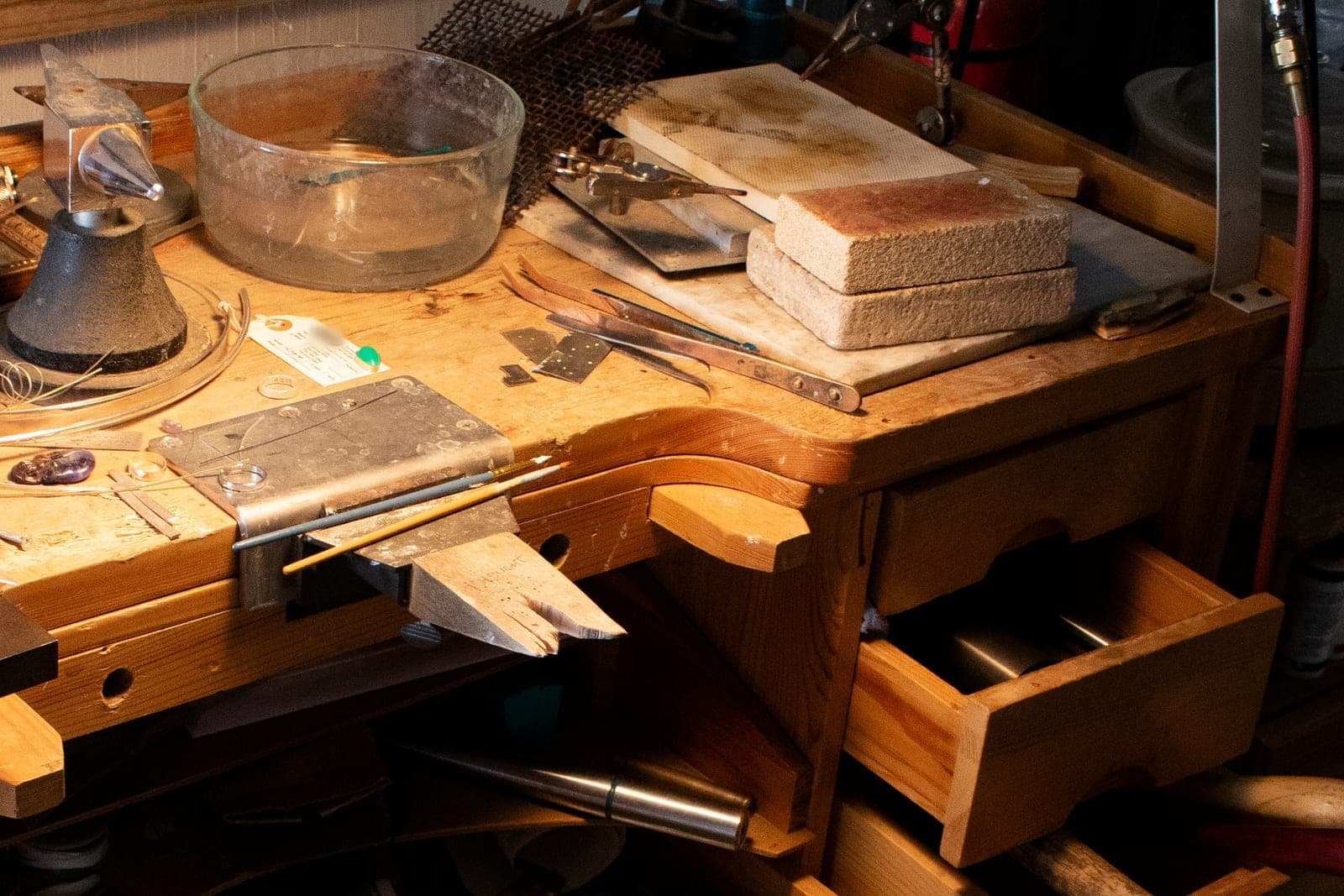 A wooden jeweler's bench holds a glass bowl, a steel horn anvil, soldering blocks, a bench pin and a stone resting on a job tag