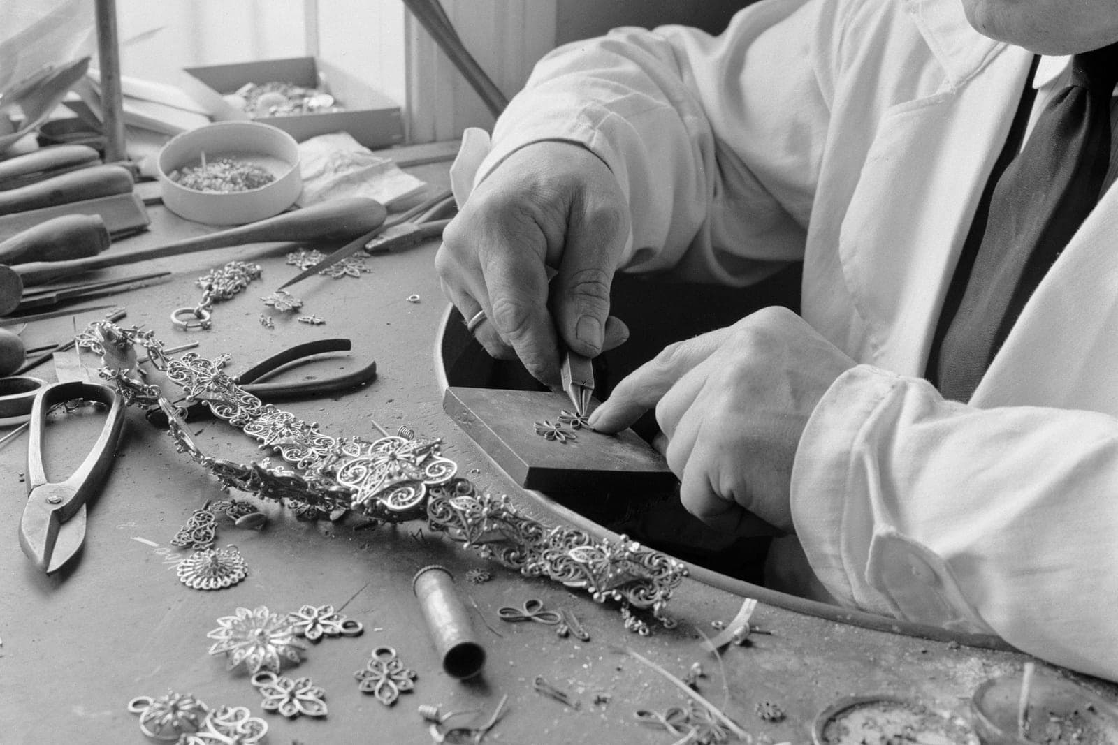 A jeweler's hands work a small silver flower with pliers on a bench covered in filigree pieces and tools