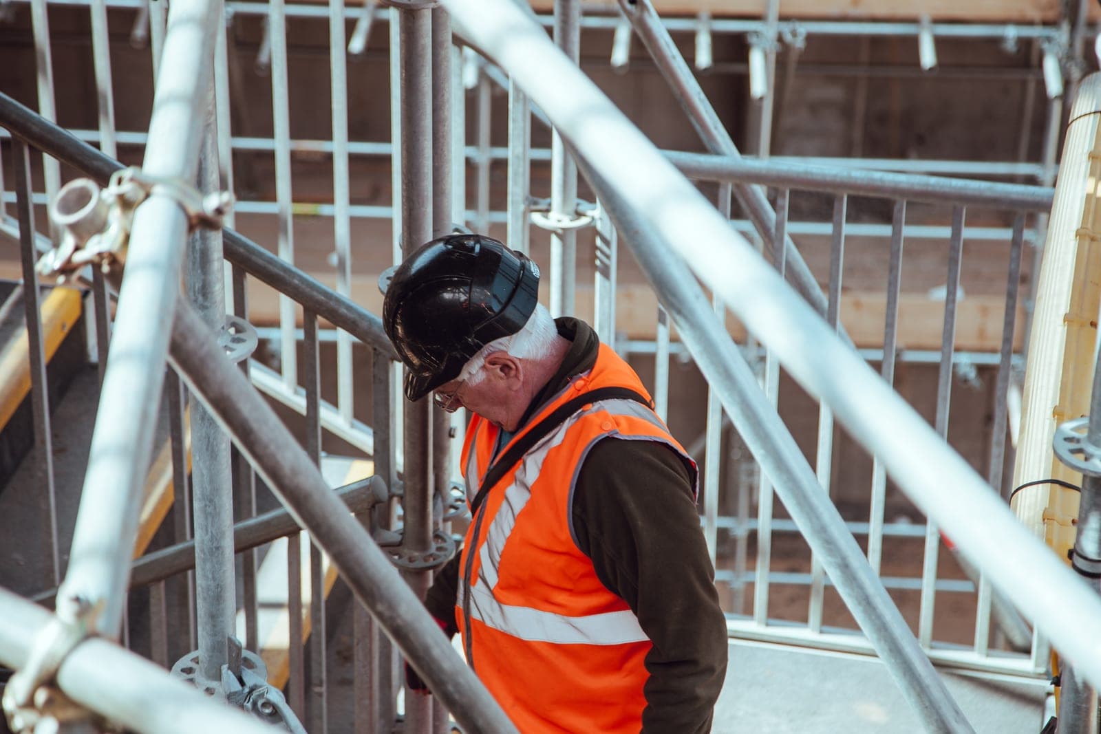 A worker in a hard hat and orange safety vest walks down scaffolding stairs on a building site