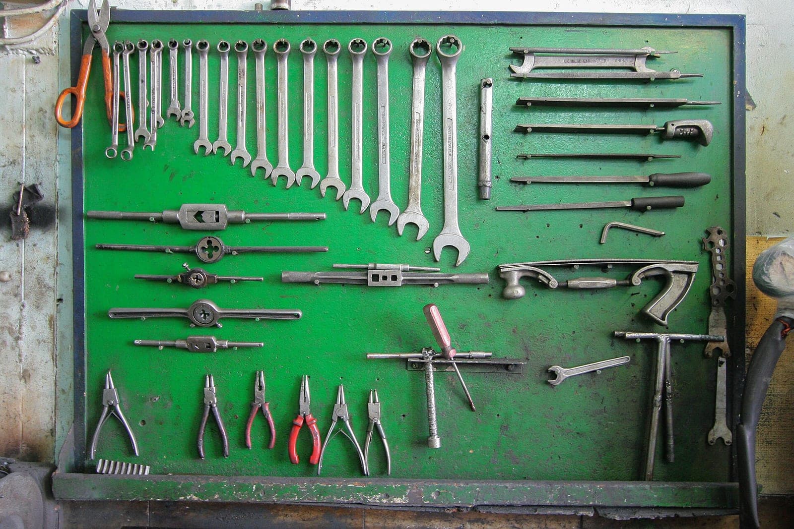 A green tool board in a machine shop holding rows of wrenches, pliers, tap handles and files