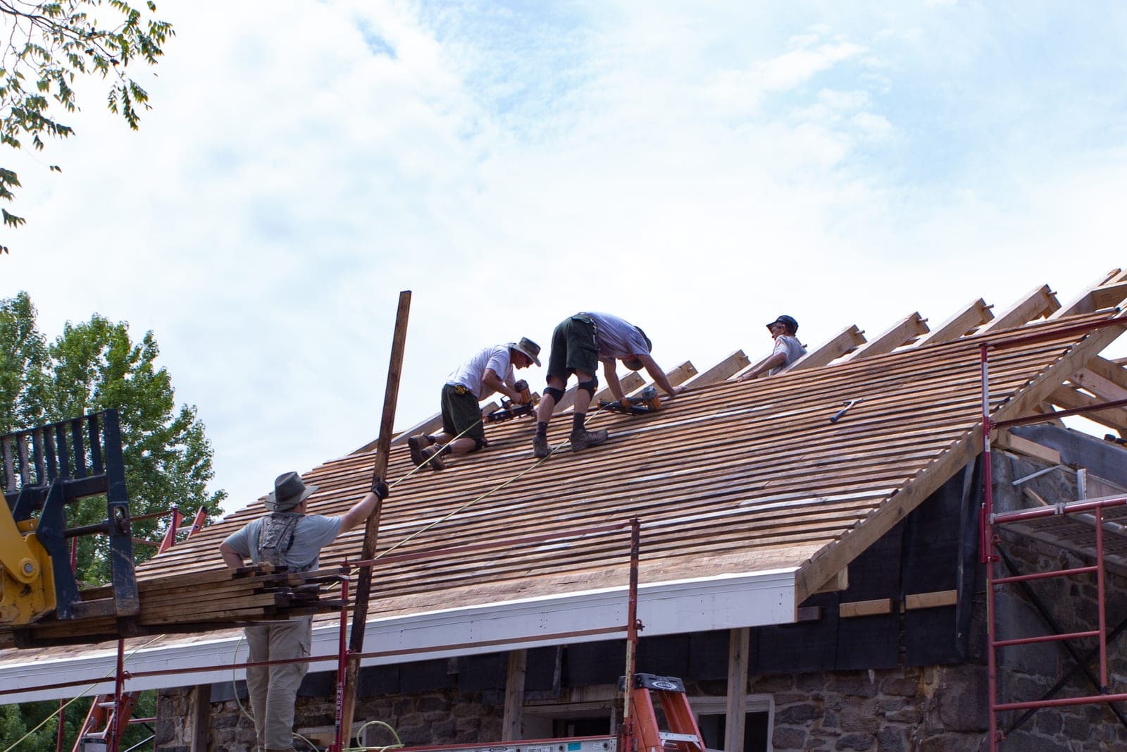 A roofing crew kneels on a steep roof laying wood strips for new shingles, while a worker on scaffolding below lifts boards