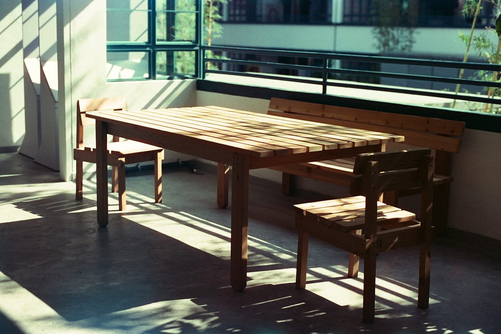 A wooden table, two chairs and a bench sit in afternoon sun on a concrete floor