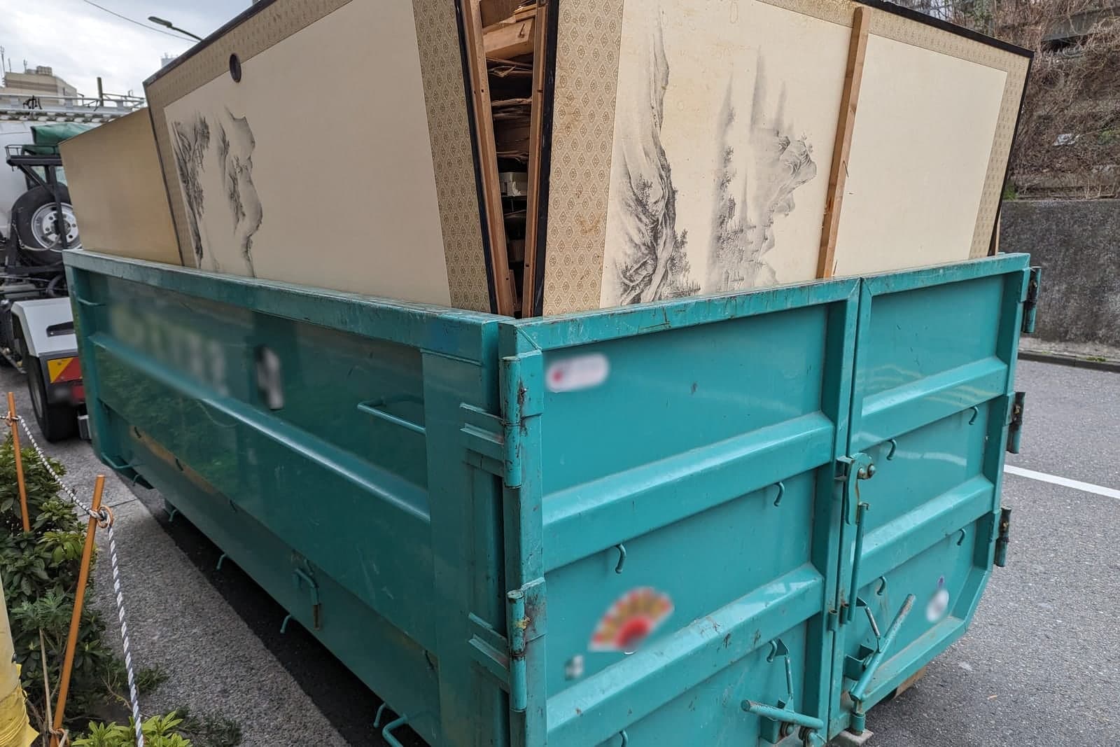 A green roll-off container at the curb is loaded with old wooden panels and doors for hauling away