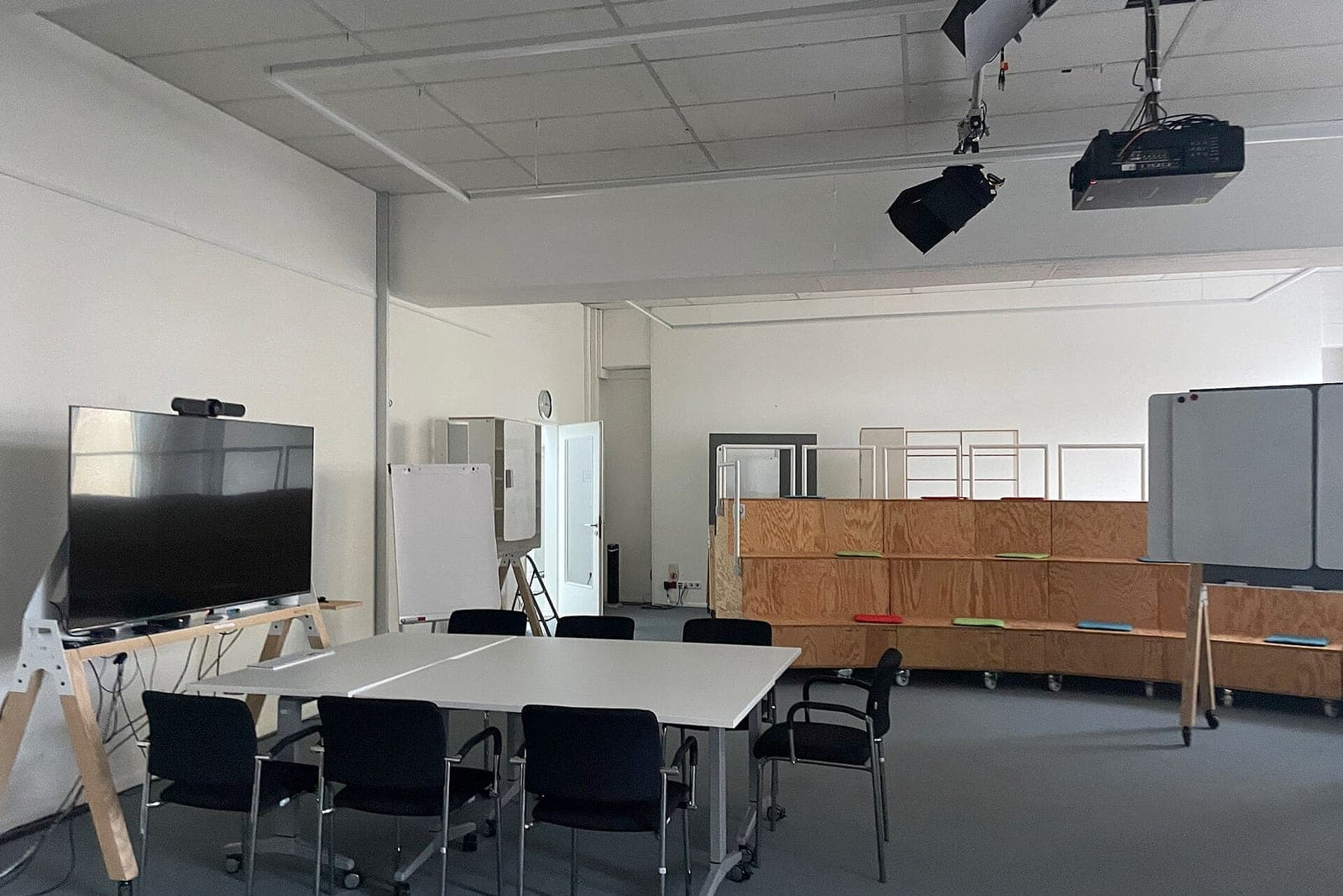 A plain meeting room with white tables, black chairs, a wall screen, a flip chart and wooden bench seating