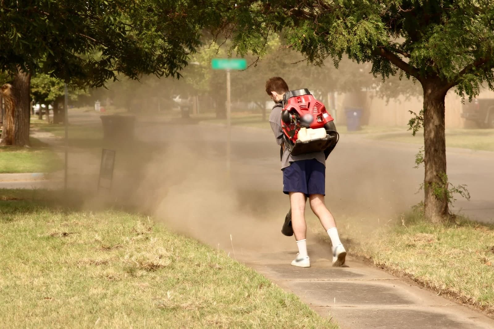A lawn care worker with a backpack blower clears grass clippings off a sidewalk after a mow