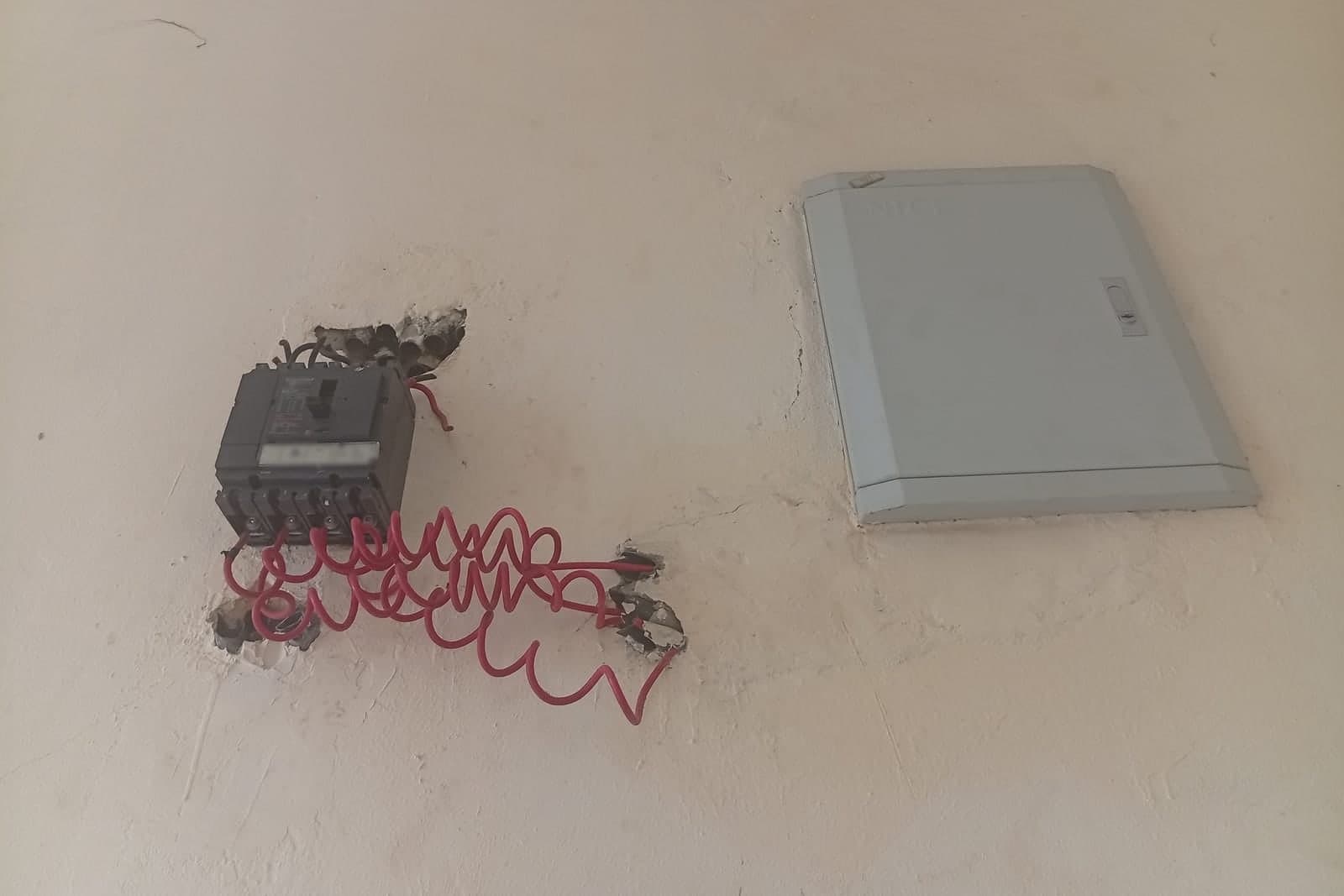 A breaker box on a plaster wall next to an old switch with loose red wiring hanging below it
