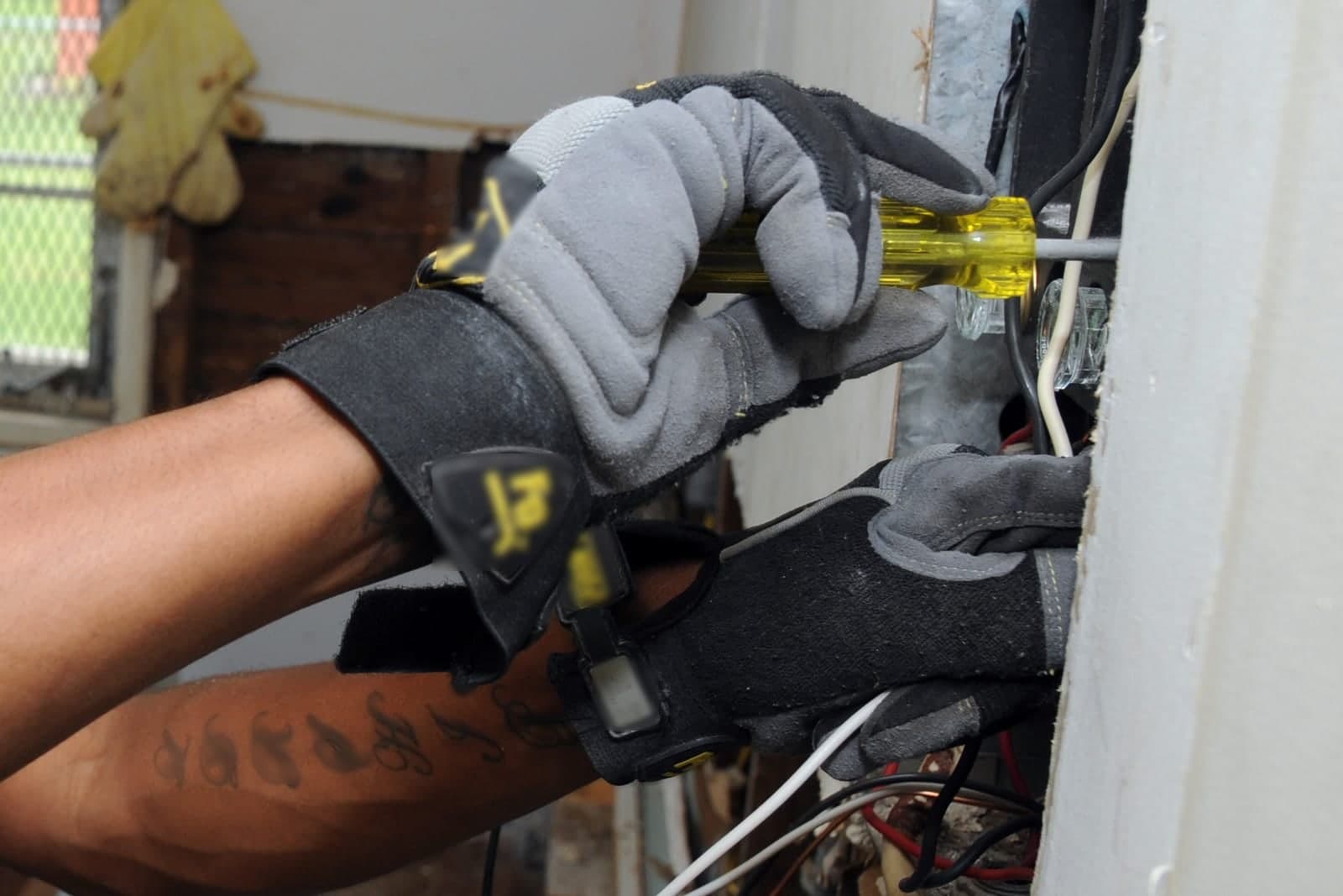 An electrician's gloved hands work on the wiring of an old fuse box inside a home