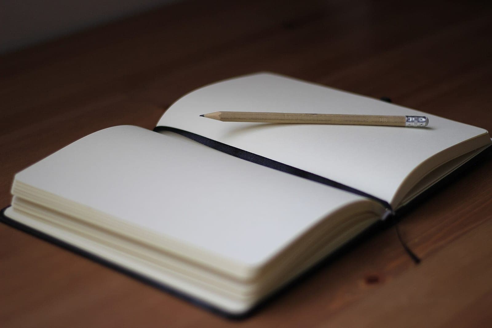 An open notebook with blank cream pages and a pencil lying across it on a wooden table