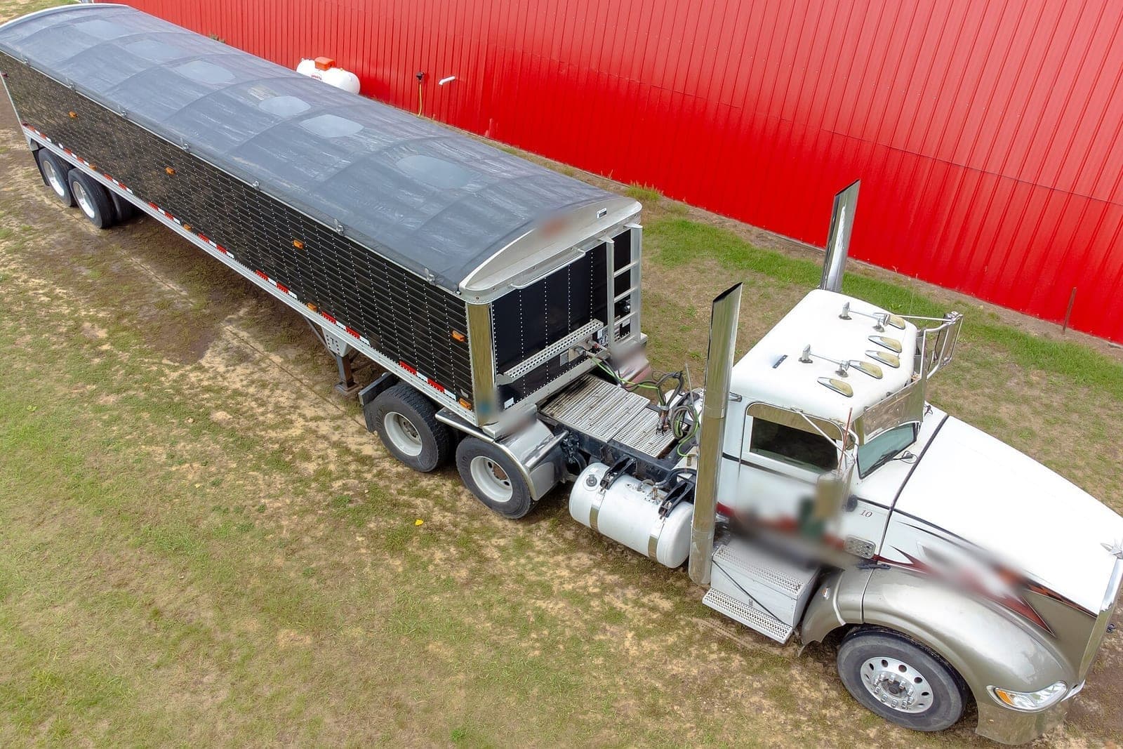 An aerial view of a white semi truck hitched to a black grain hopper trailer, parked on grass beside a red metal building