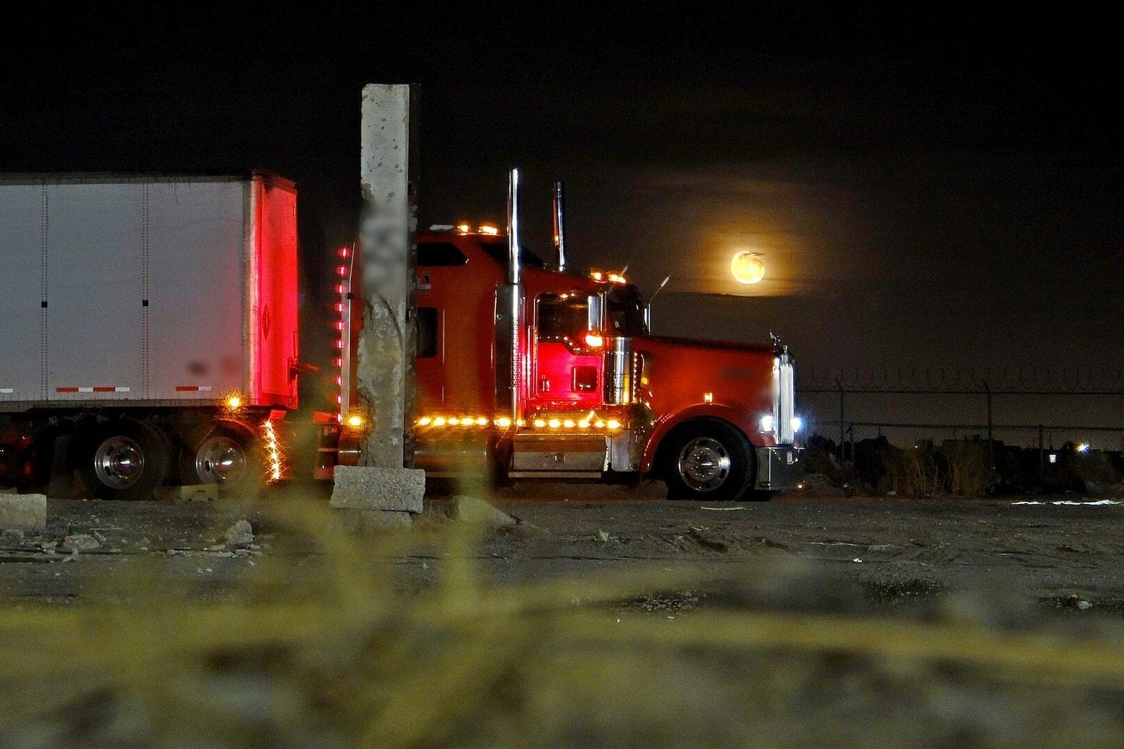 A red semi truck with glowing marker lights sits parked beside a white trailer at night under a rising full moon