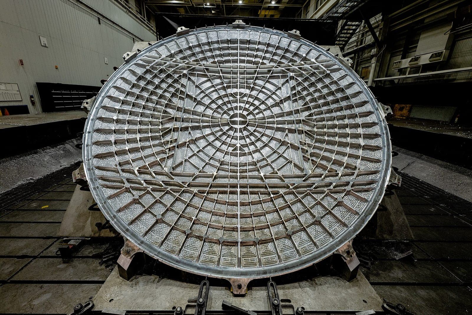 A large round aluminum part with a ribbed grid pattern sits clamped on the bed of a big milling machine