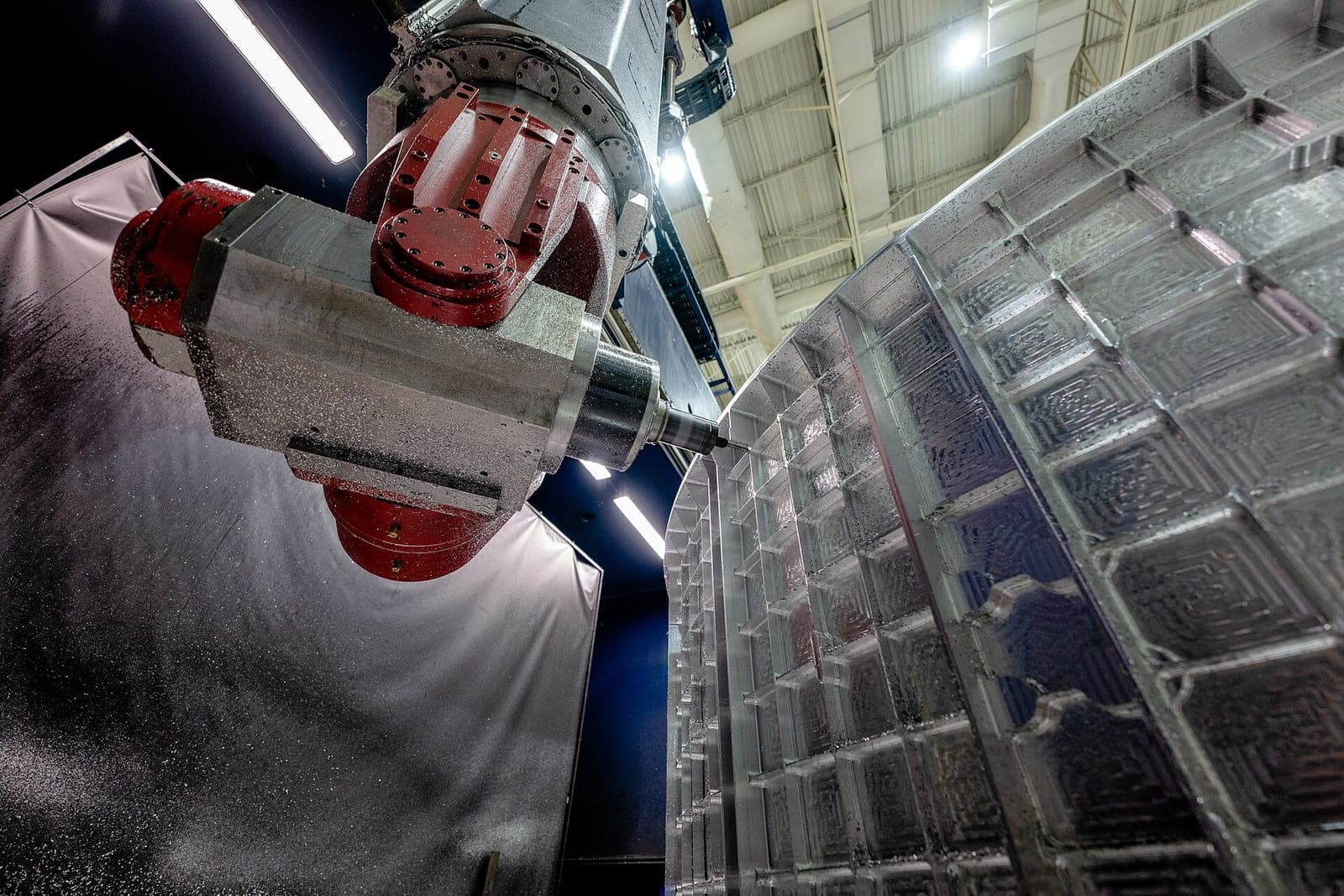 A red milling head cuts a waffle pattern into a large curved aluminum part inside a machine shop