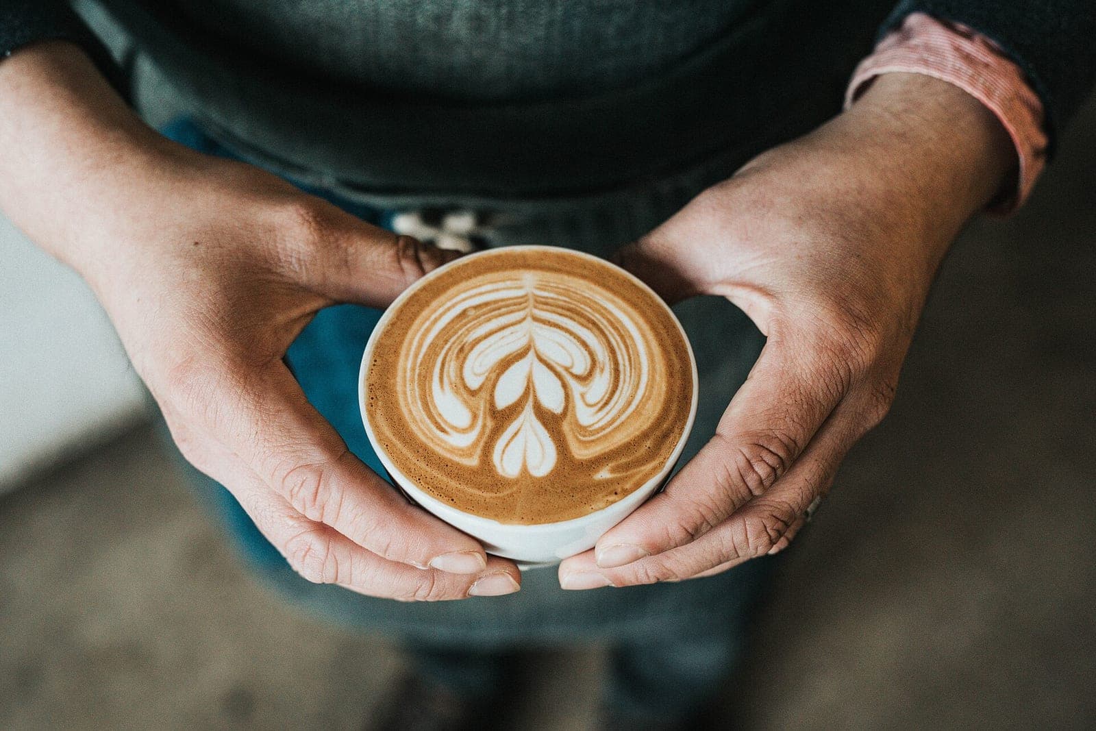 Two hands holding out a white cup of coffee with leaf-pattern latte art