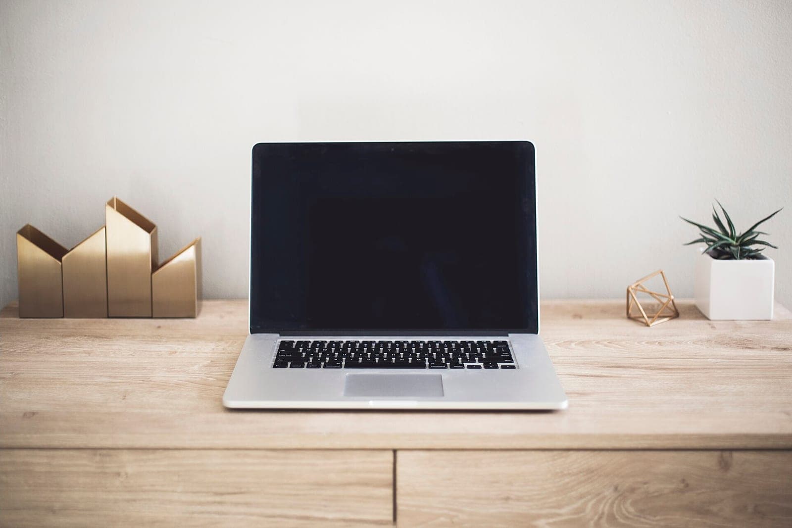A laptop with a dark screen on a light wood counter between gold desk pieces and a small potted plant