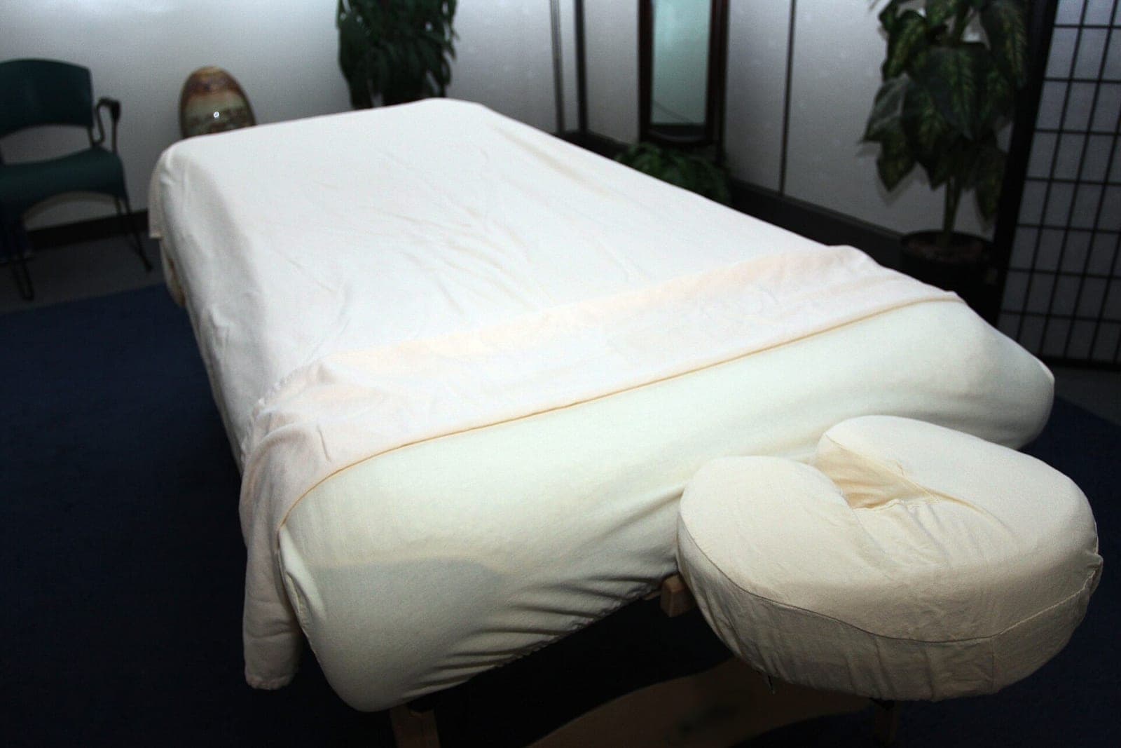 An empty treatment room with a padded table made up in cream sheets, a face rest at one end and potted plants behind it