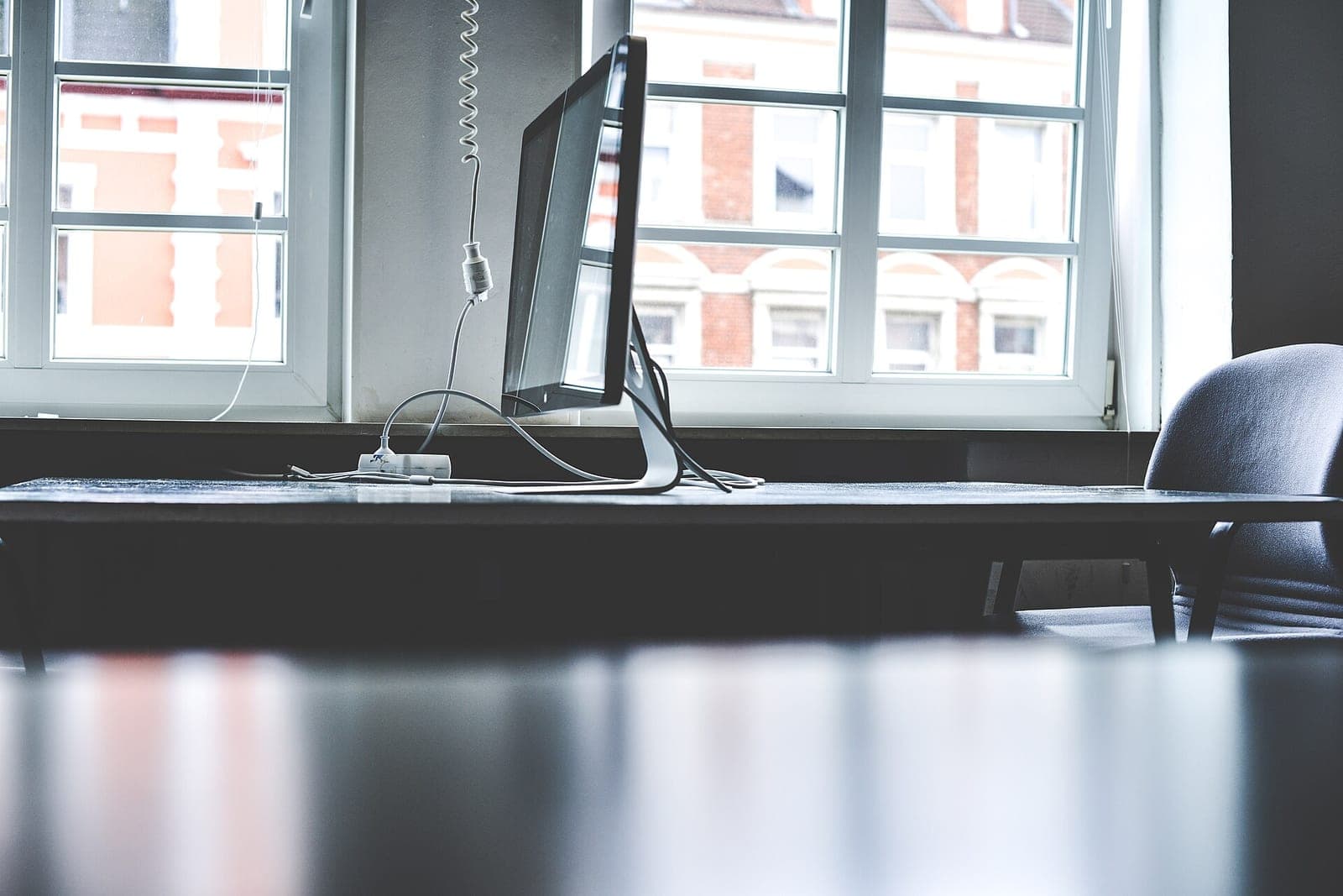 A computer monitor on a dark desk in front of tall white-framed windows, with an office chair to one side