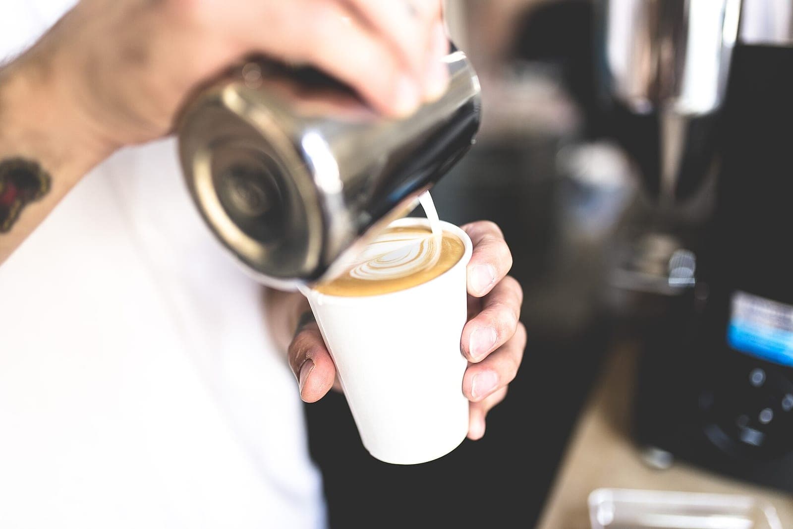 A barista's hands pour steamed milk from a metal pitcher into a white paper cup, making a leaf pattern