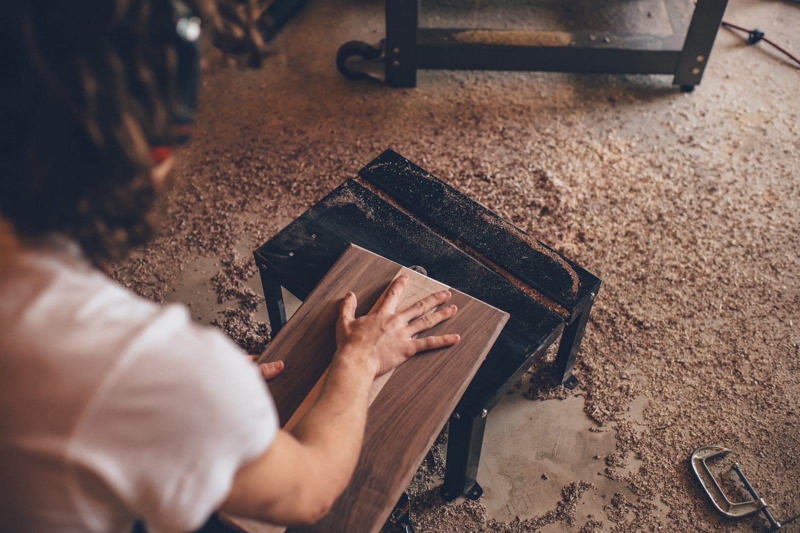 A woodworker, seen from behind, presses a dark wooden board flat on a small bench on a shop floor covered with sawdust