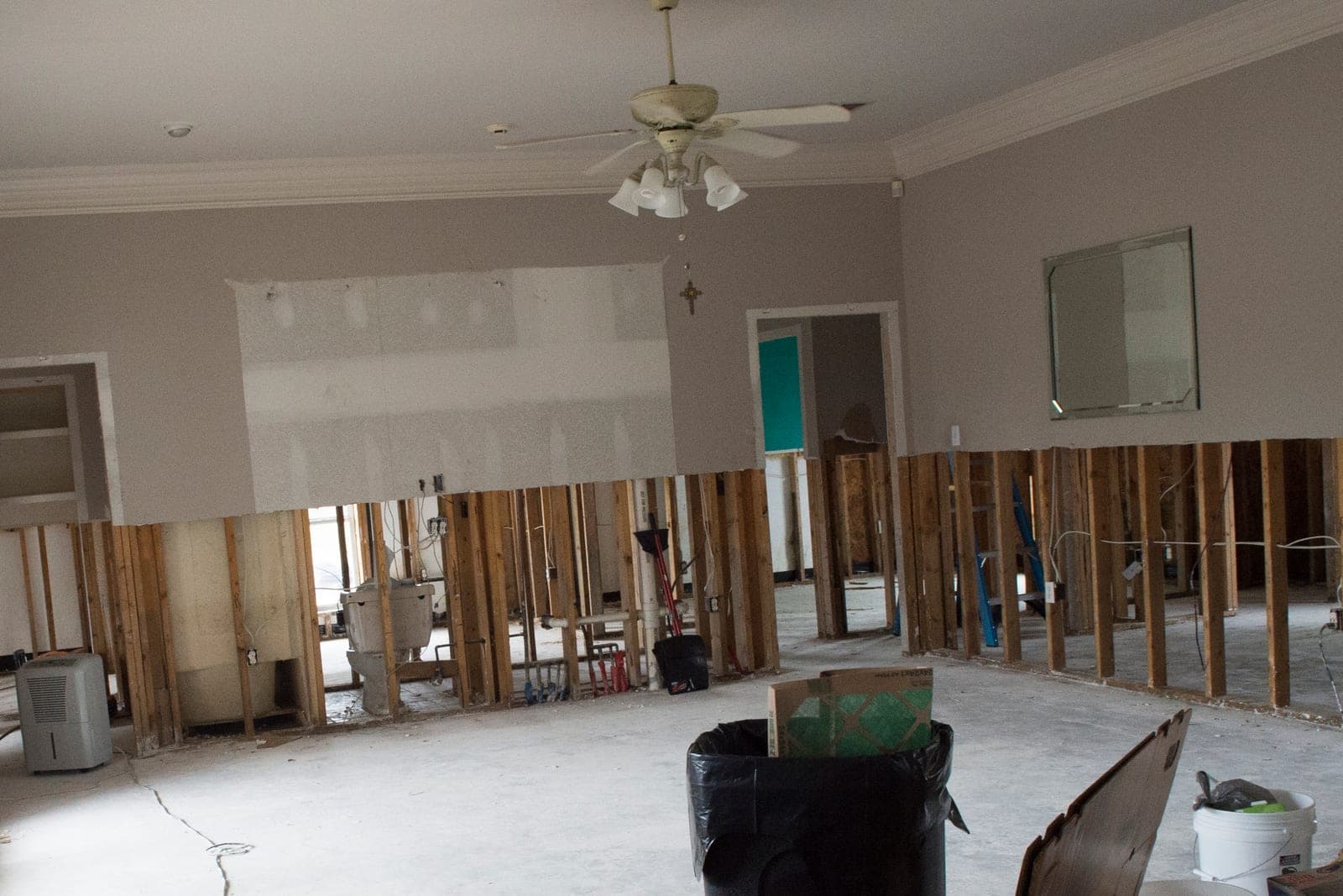 Wet drywall has been cut away along the lower walls of an empty living room, leaving bare studs and a small dehumidifier in the corner