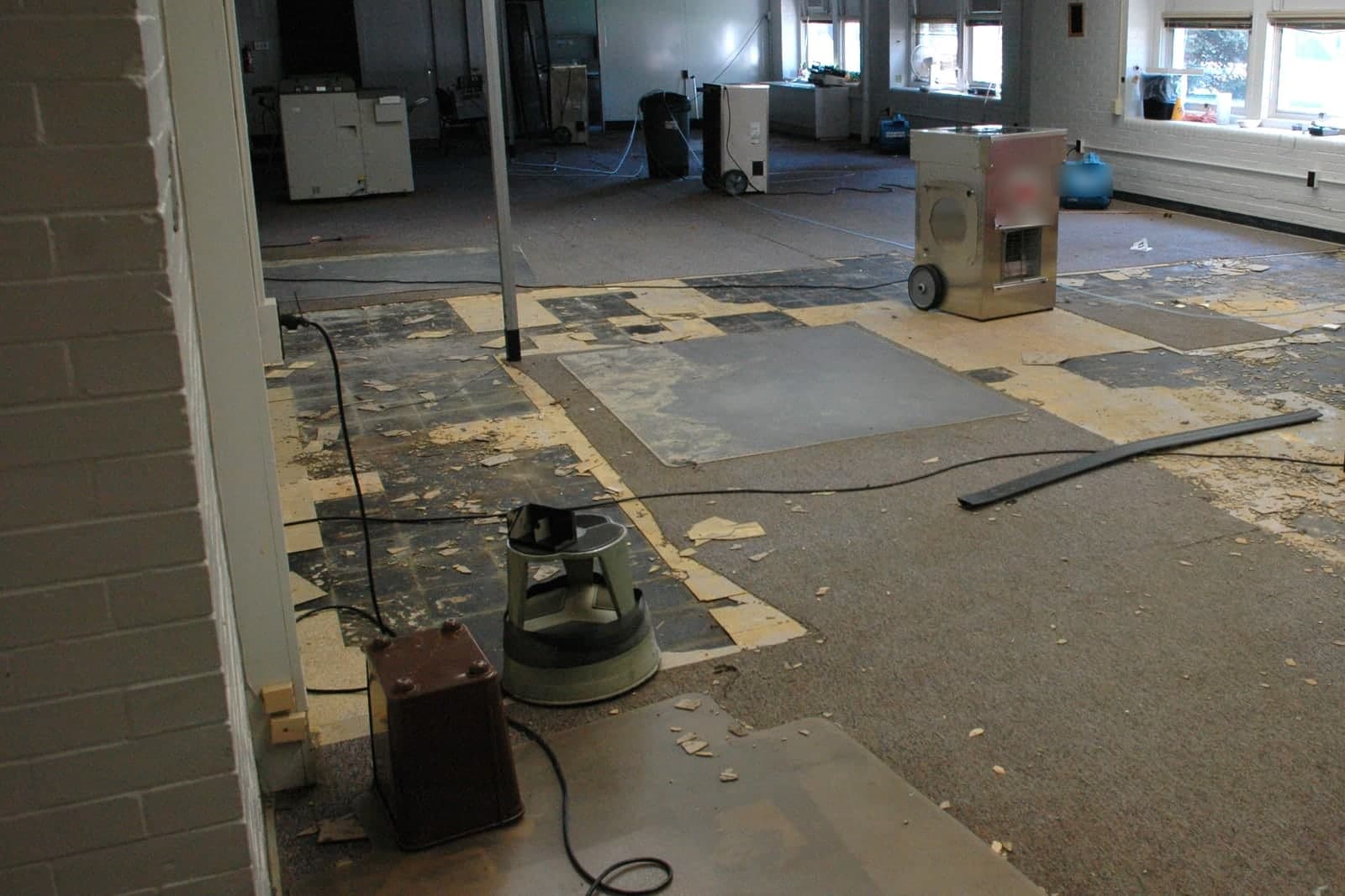 A steel dehumidifier and a blue air mover sit on a half-stripped tile floor in an empty room, with power cords running across it