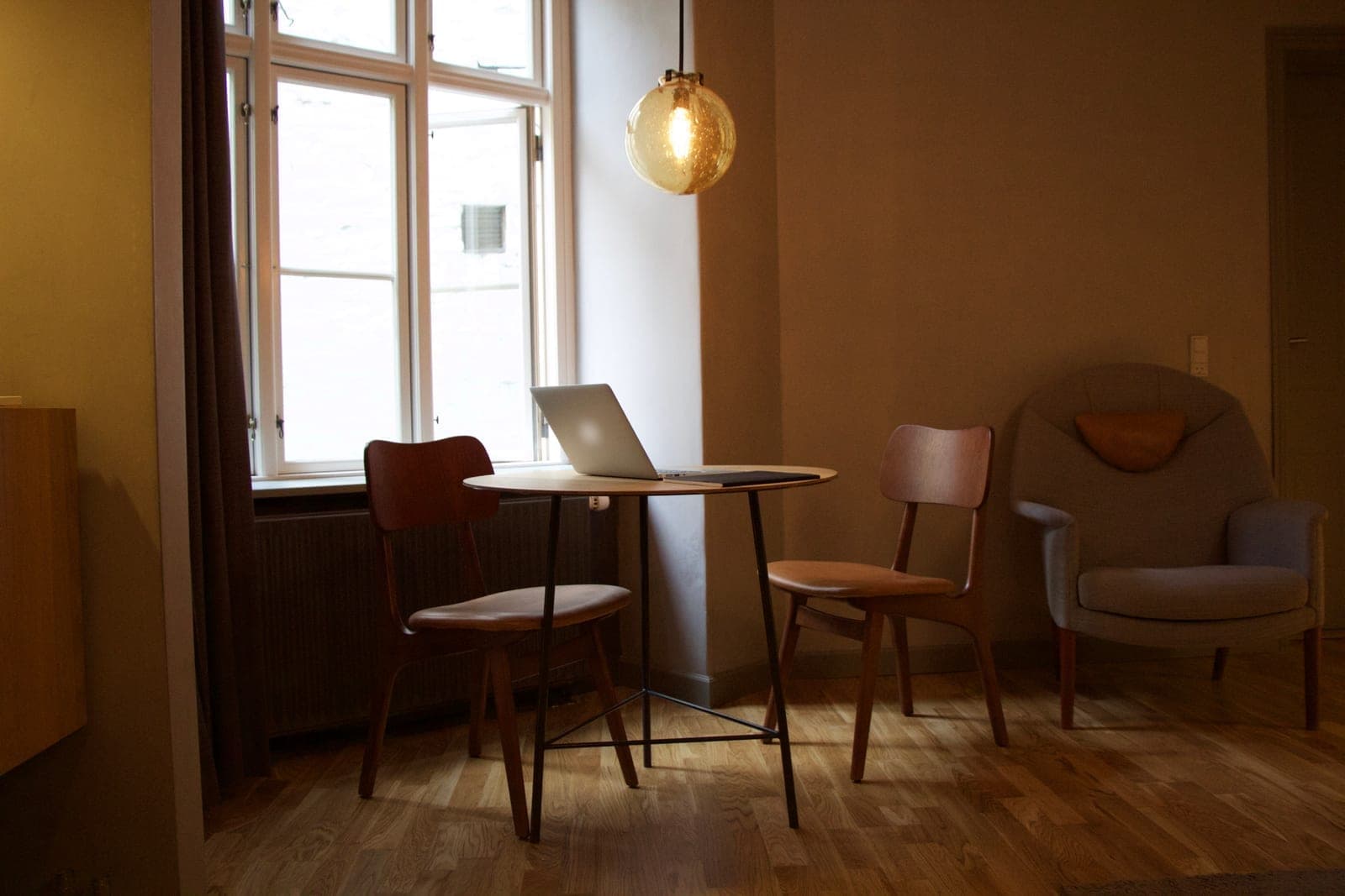 A laptop on a small round table by a window in a quiet living room, with two wooden chairs and a gray armchair