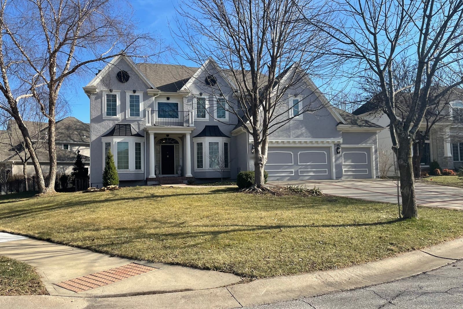 A gray two-story house with a front balcony, a three-car garage and bare trees on a sunny winter day in Leawood, Kansas