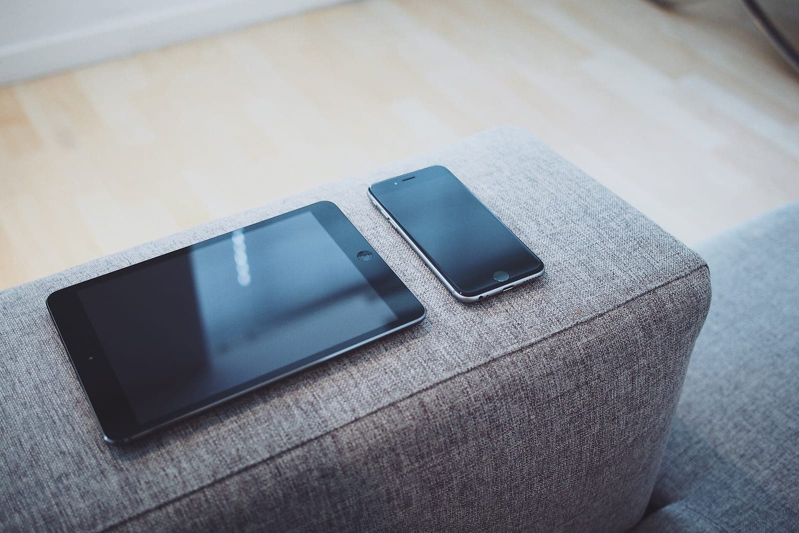 A tablet and a phone resting side by side on the arm of a gray sofa