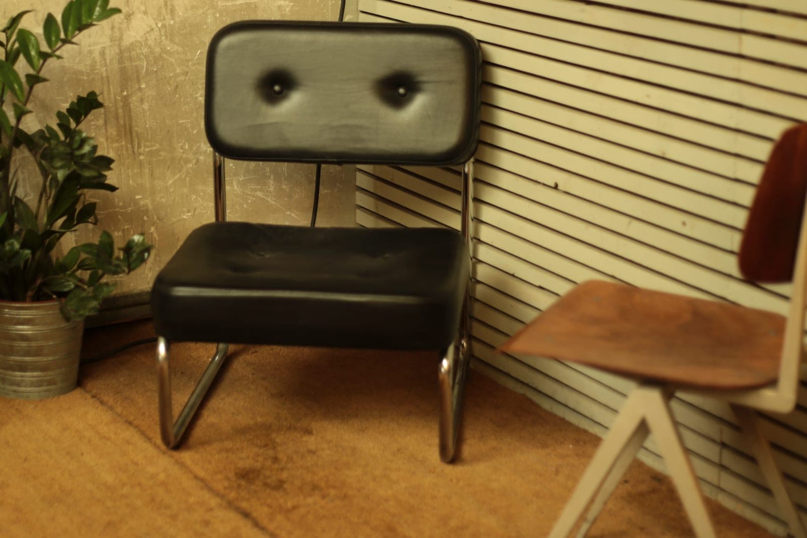 A black padded chair on a chrome frame stands in a room corner beside a potted plant and a wooden chair