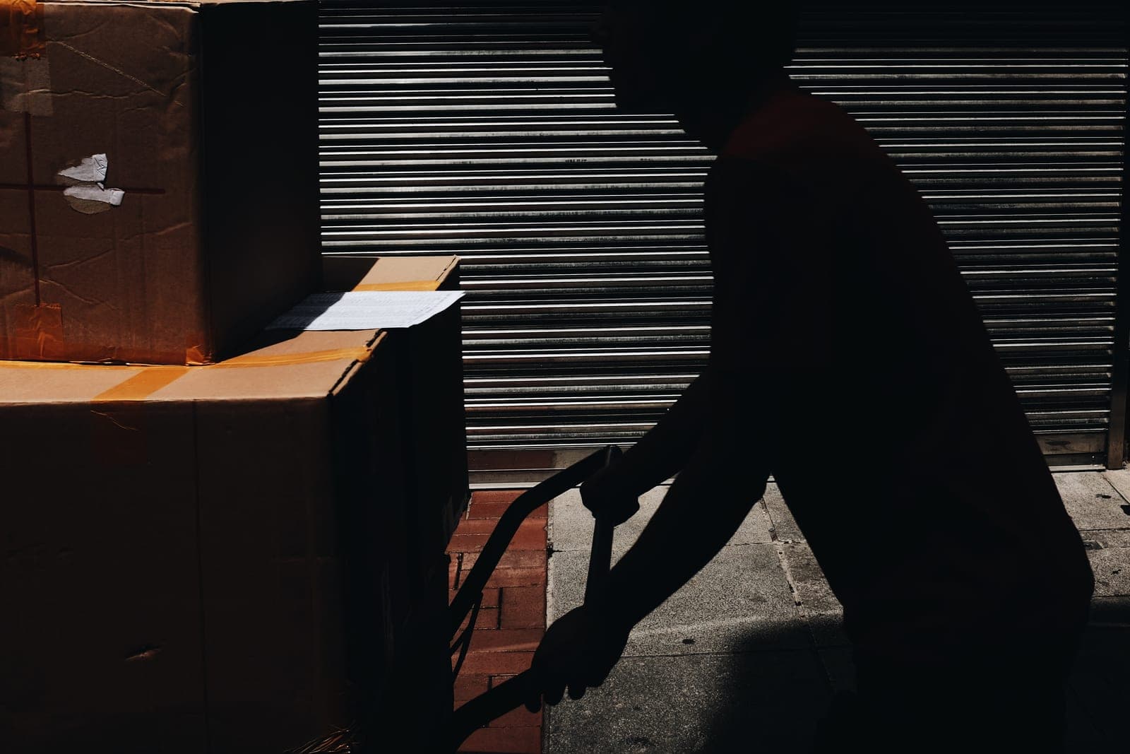 A person in silhouette pushes a hand truck stacked with large cardboard cartons past a closed metal shutter