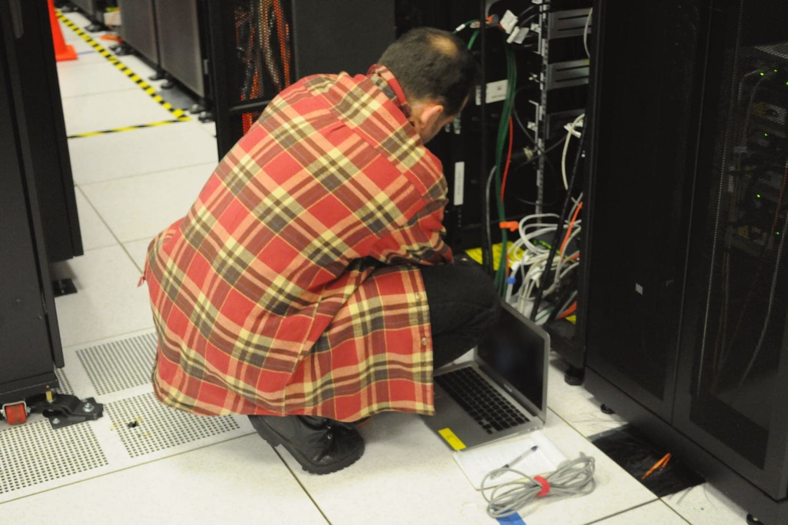 A technician in a plaid shirt crouches at an open server rack with a laptop on the floor