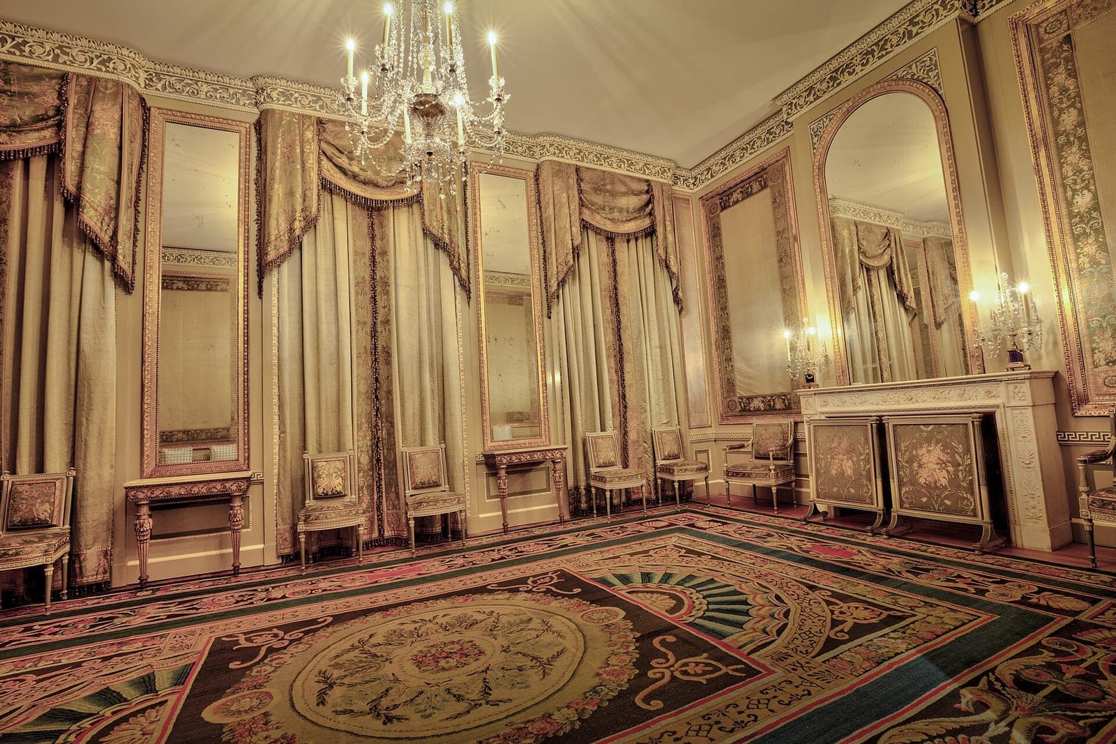 An empty formal salon with tall mirrors, silk drapes, gilded chairs along the walls and a patterned carpet under a crystal chandelier
