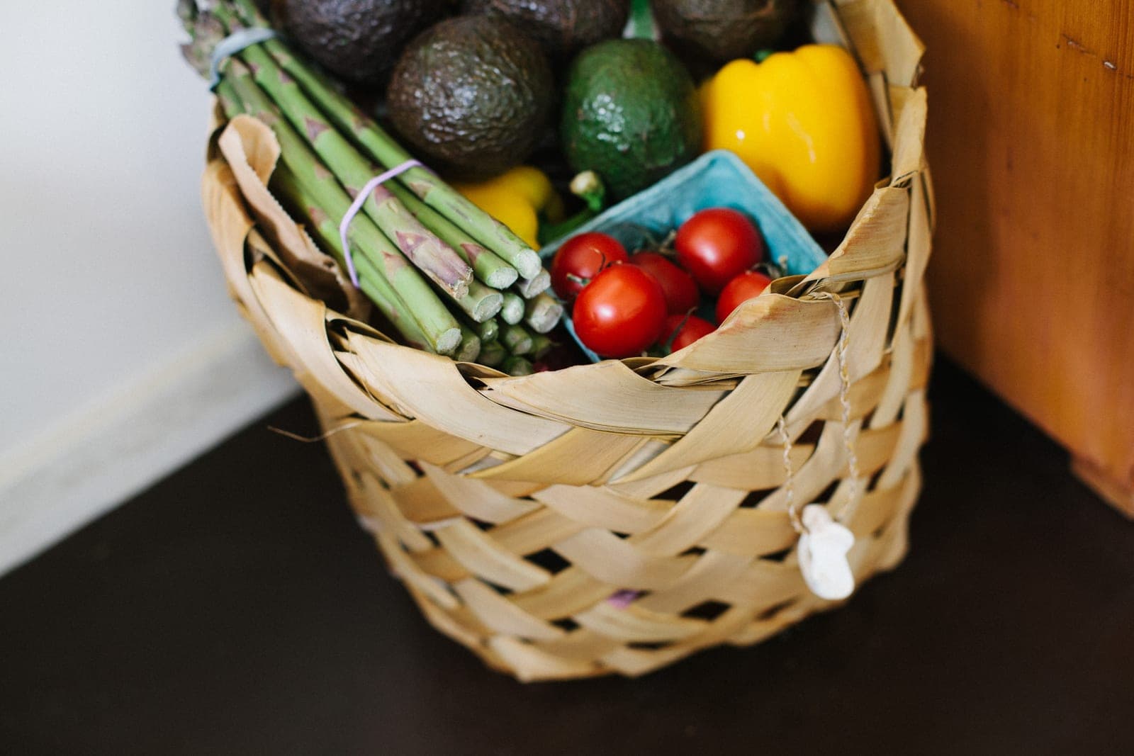 A woven basket holds asparagus, avocados, yellow peppers and a carton of small tomatoes on a dark floor