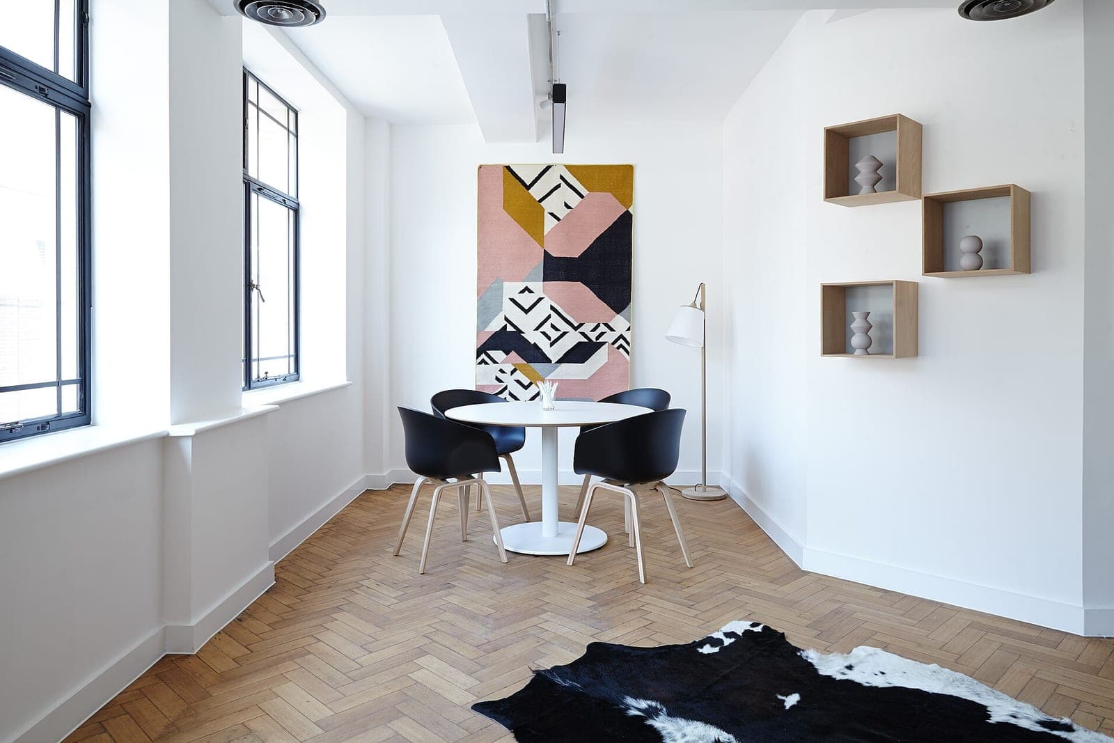 A white dining nook with a round table, four black chairs, wall shelves and a geometric wall hanging