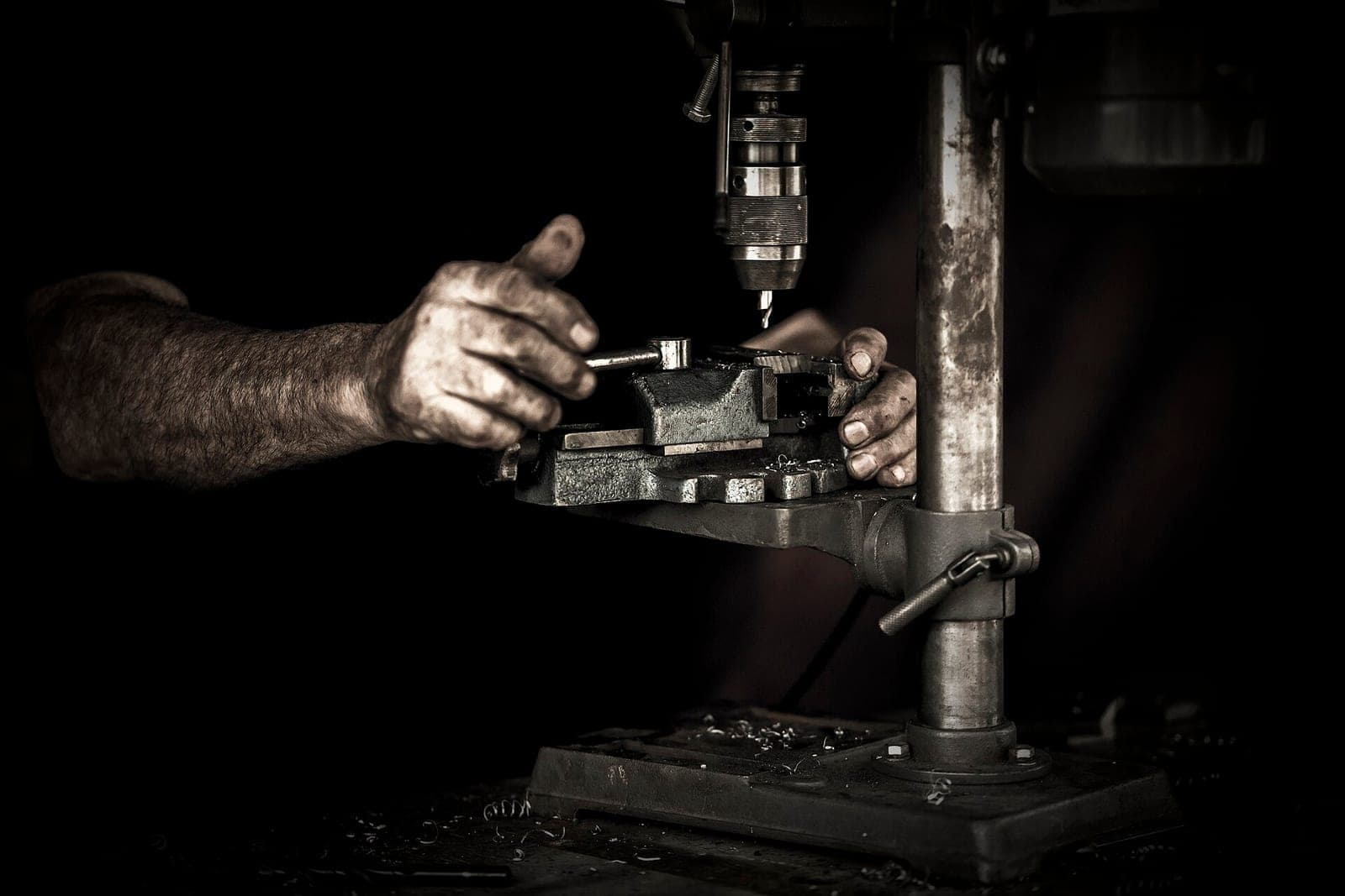 Two work-worn hands steady a metal vise under a bench drill press in a dark workshop