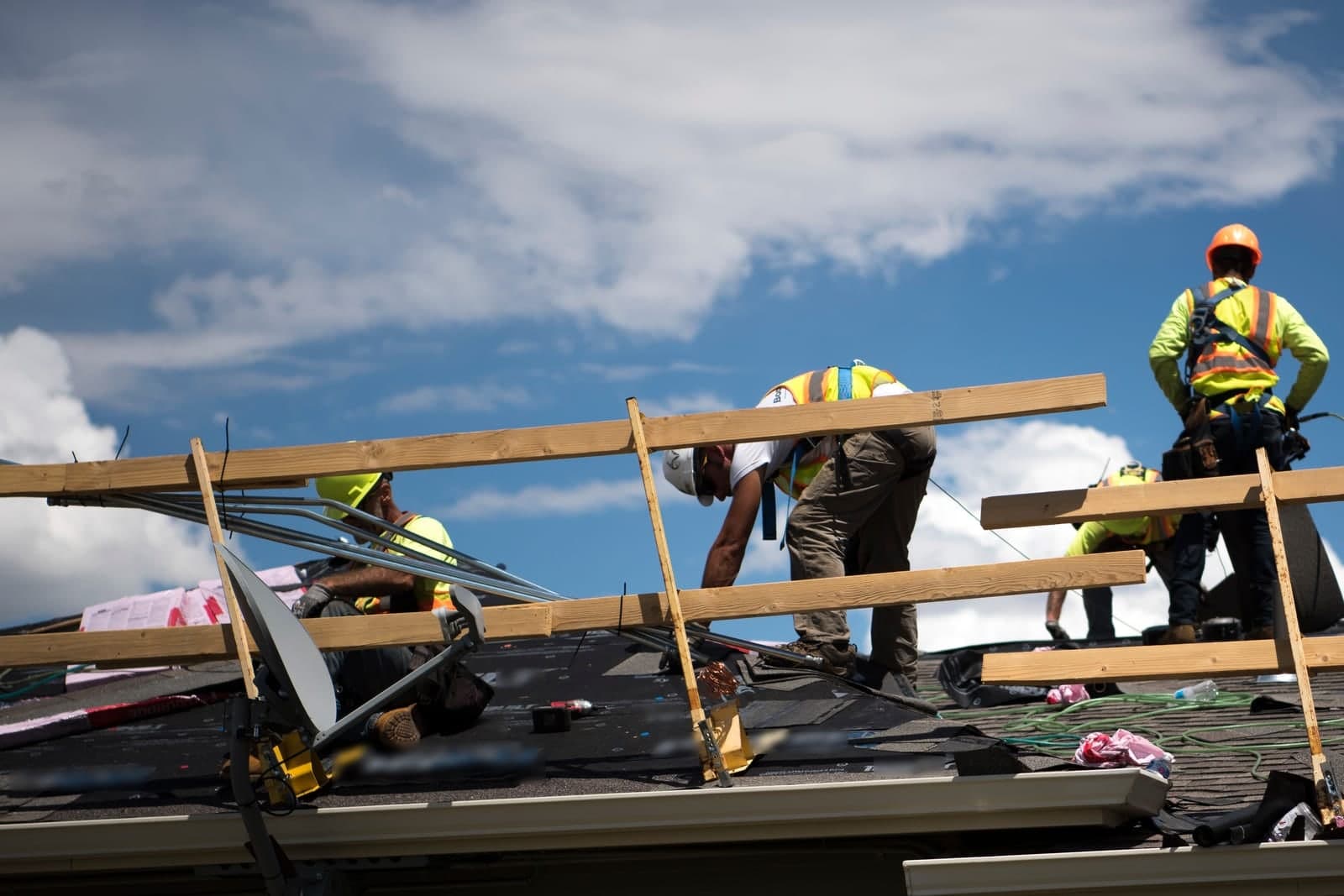 Roofers in hard hats and safety vests work on a house roof under a blue sky with white clouds