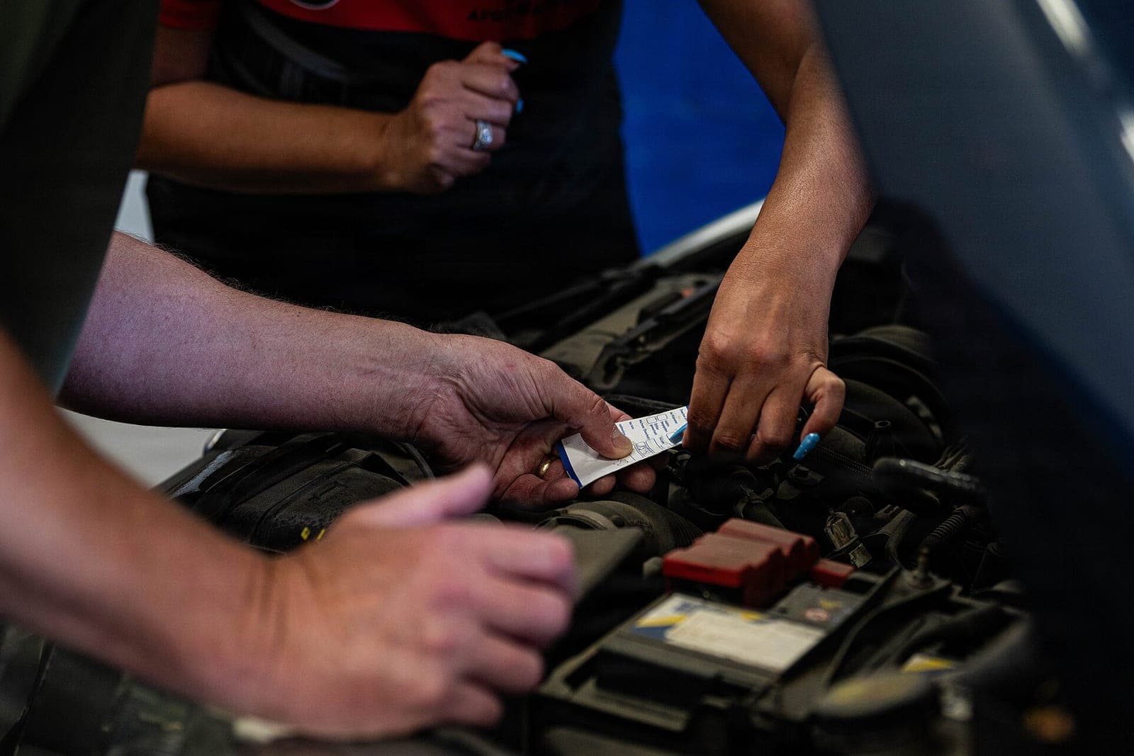 Several hands work under the open hood of a car, one of them holding a small paper tag above the battery