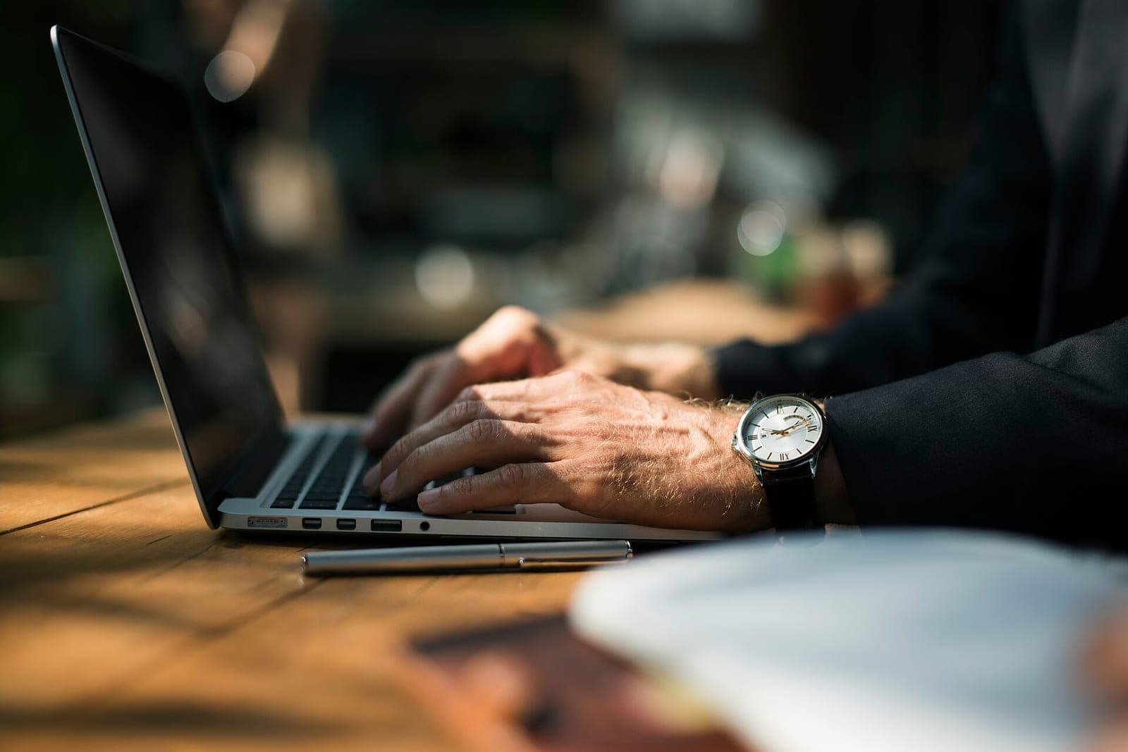 Hands typing on a laptop on a sunlit wooden table, with a silver pen beside it