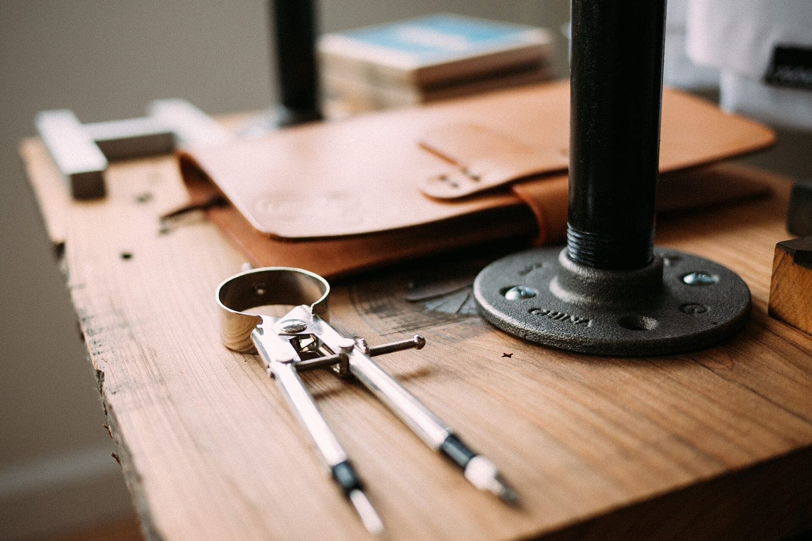 A pair of drafting dividers lies on a wooden workbench beside a tan leather folio and a black pipe stand