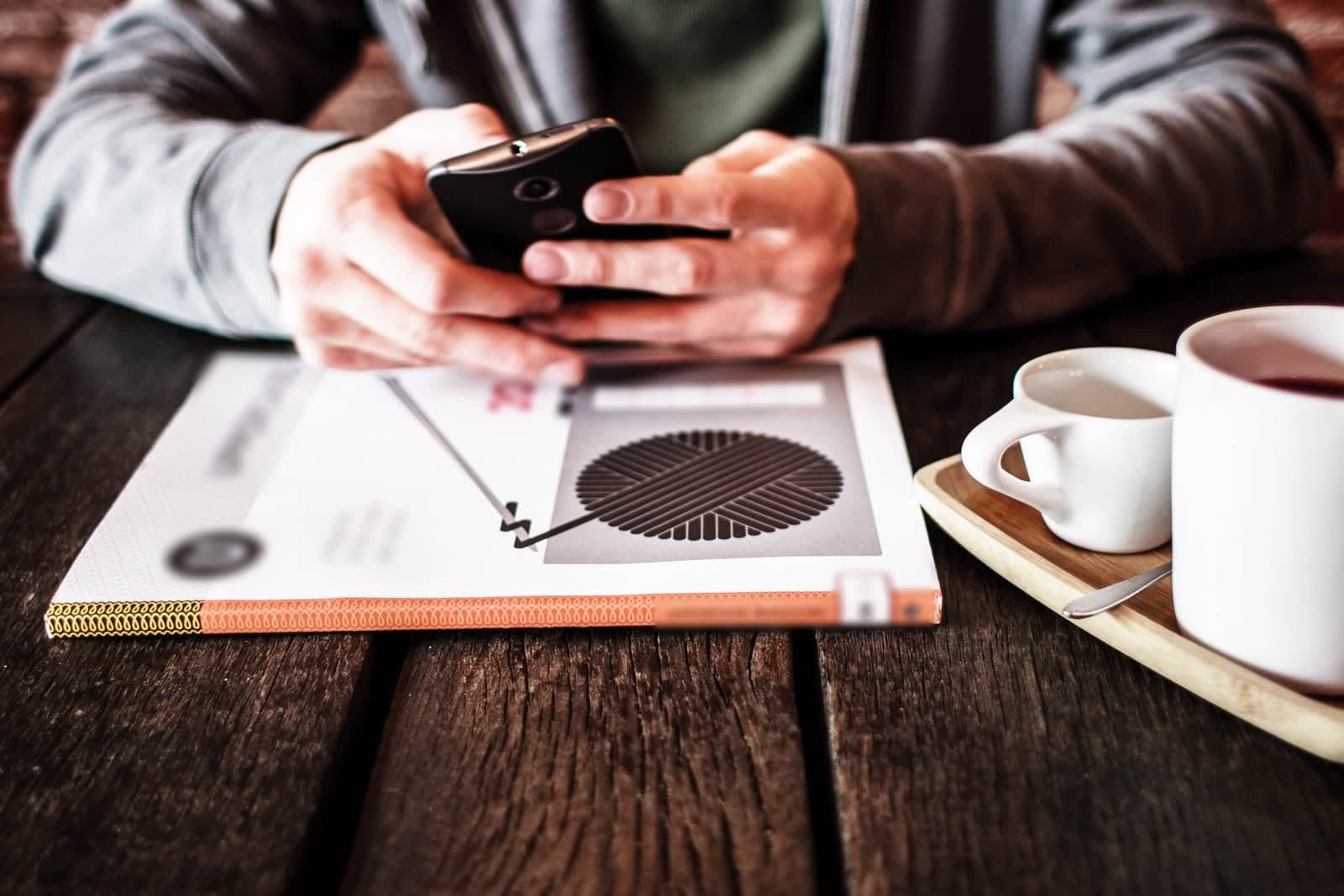 Two hands type a message on a phone above a magazine on a wooden table beside cups of coffee