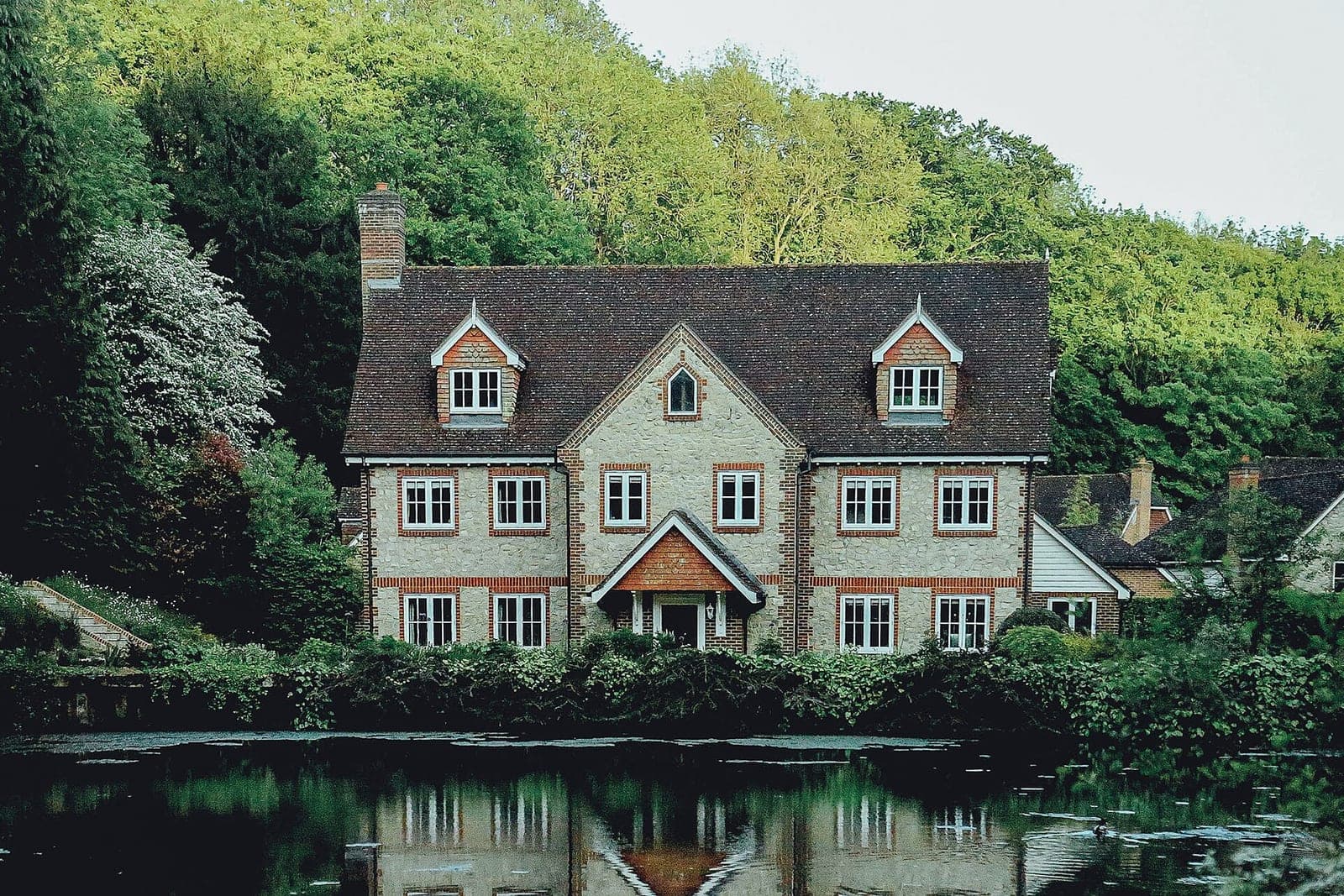 A large brick and stone house with green lawns and trees, reflected in a pond