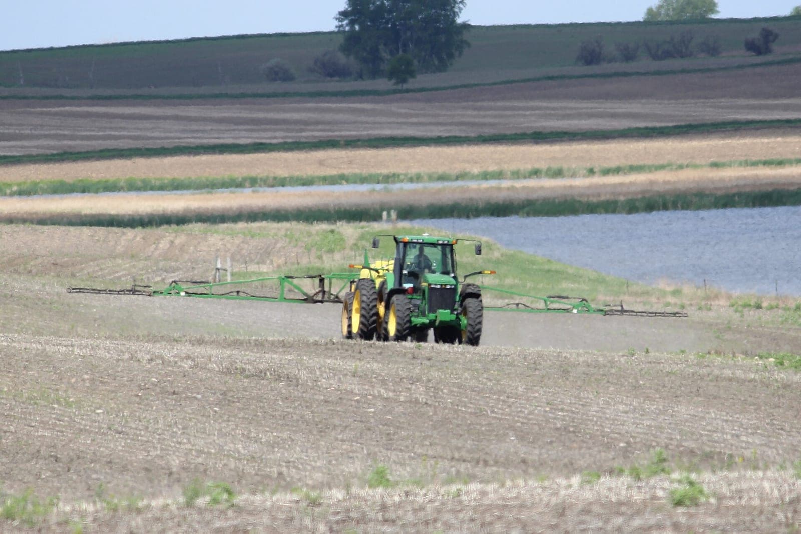 A green tractor pulls a wide boom sprayer across a bare field beside a pond