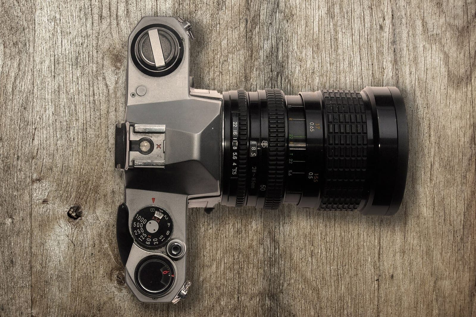 A silver film camera with a long zoom lens lies on a weathered wooden table, seen from above