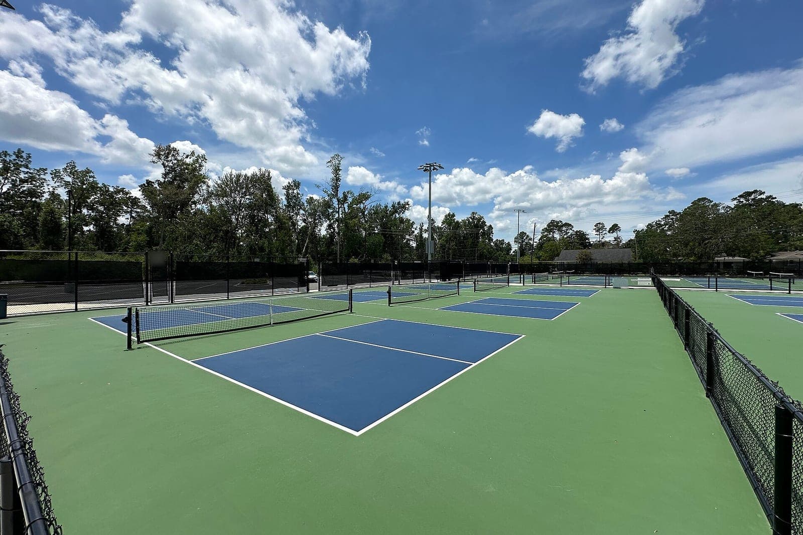 Empty outdoor pickleball courts with blue playing areas, green borders, black nets and tall light poles under a cloudy sky