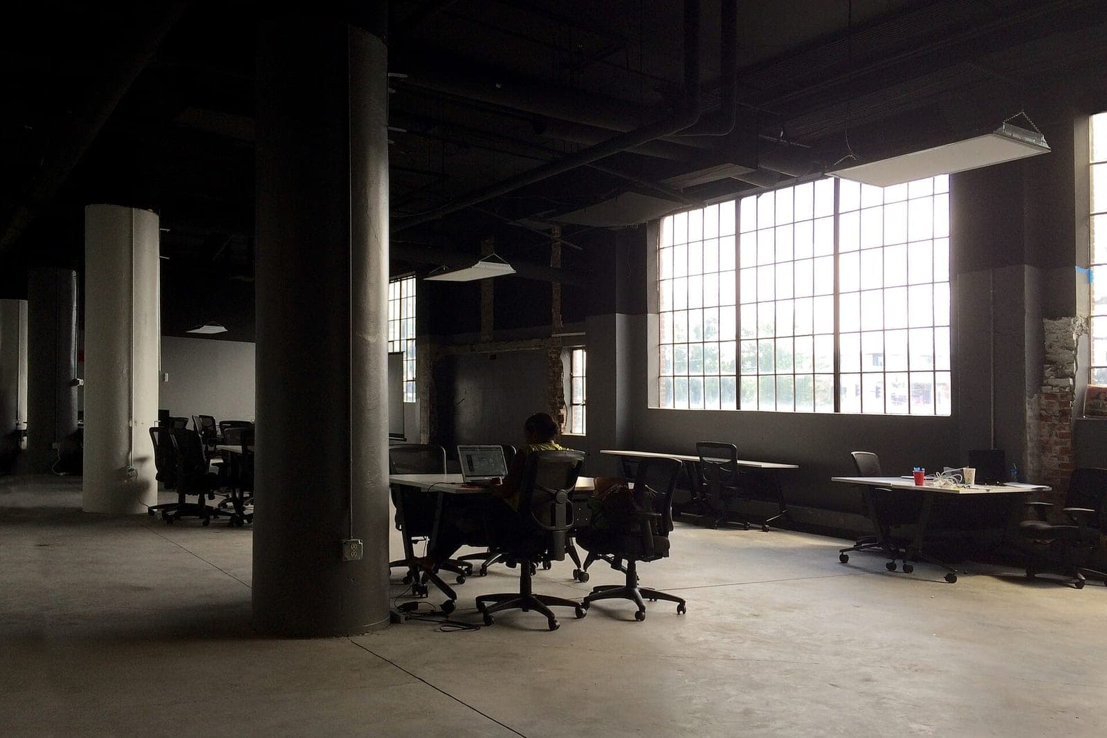 A person works at a laptop at a long table in a large open loft office with tall windows