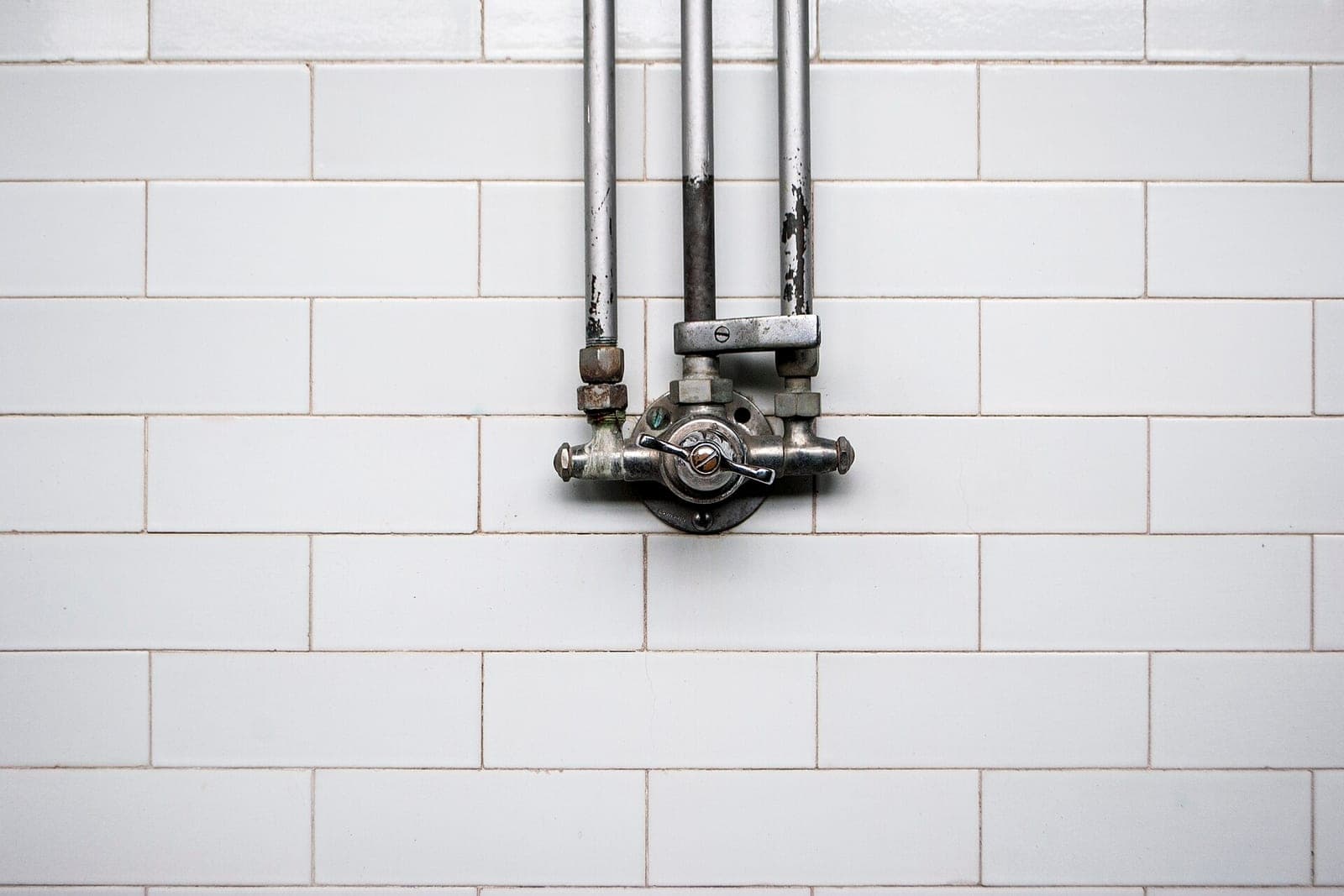 An old exposed shower valve with three metal pipes running up a white subway-tile wall