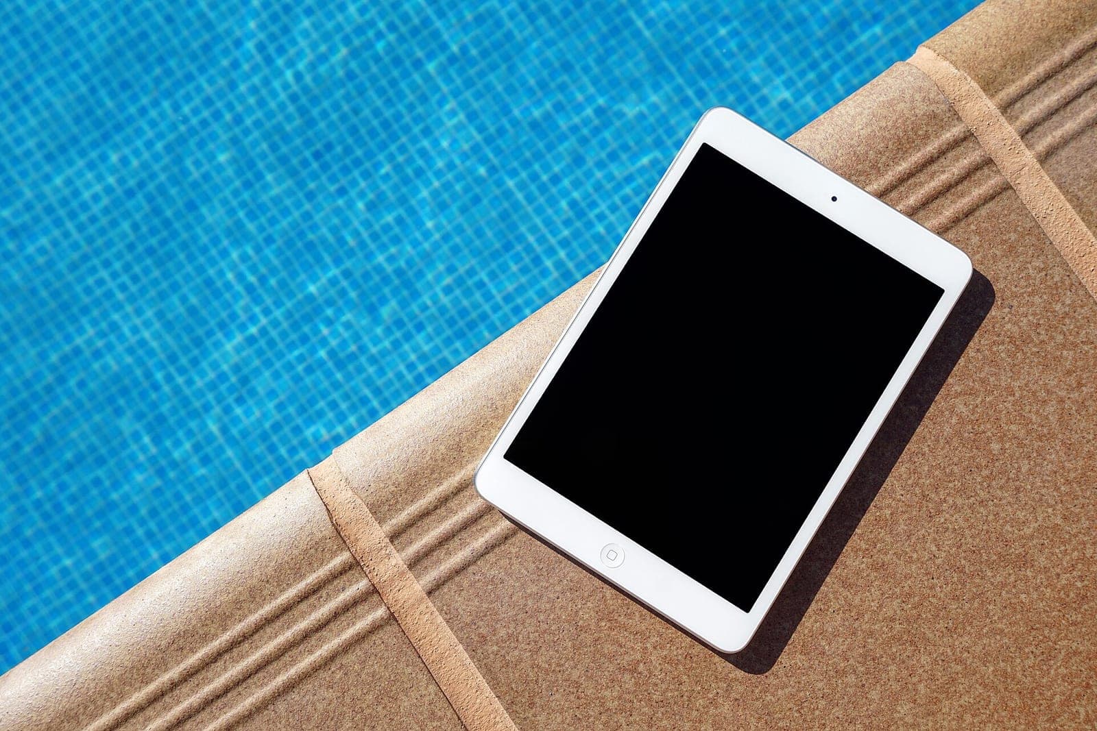 A white tablet lying on the warm stone edge of a blue tiled swimming pool