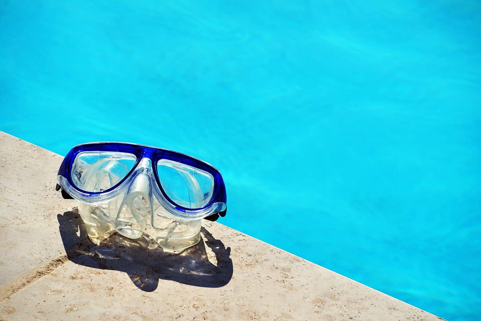 A swim mask resting on a stone pool edge beside clear blue water