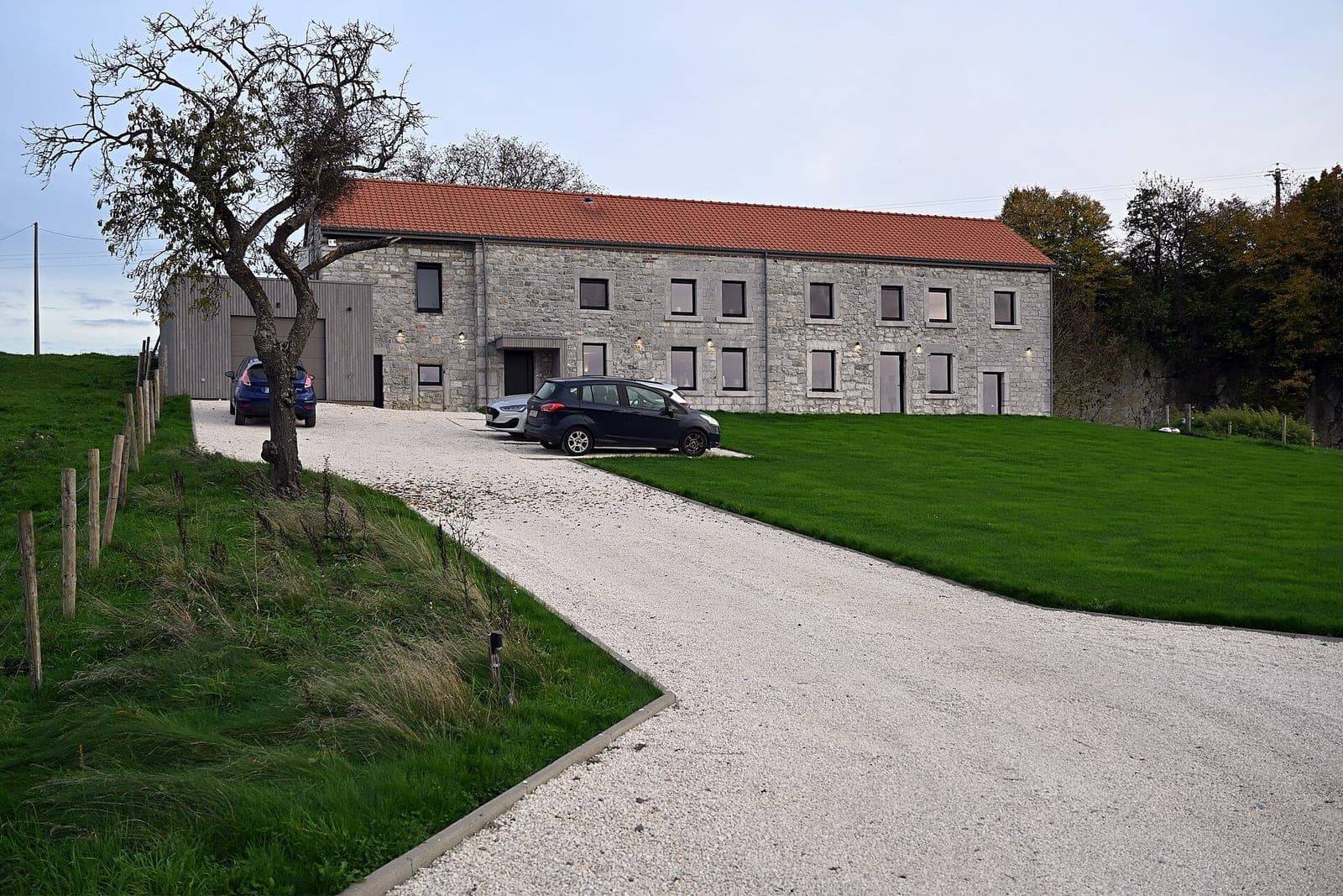 A long pale gravel driveway curving past a green lawn to a stone house with a red tile roof